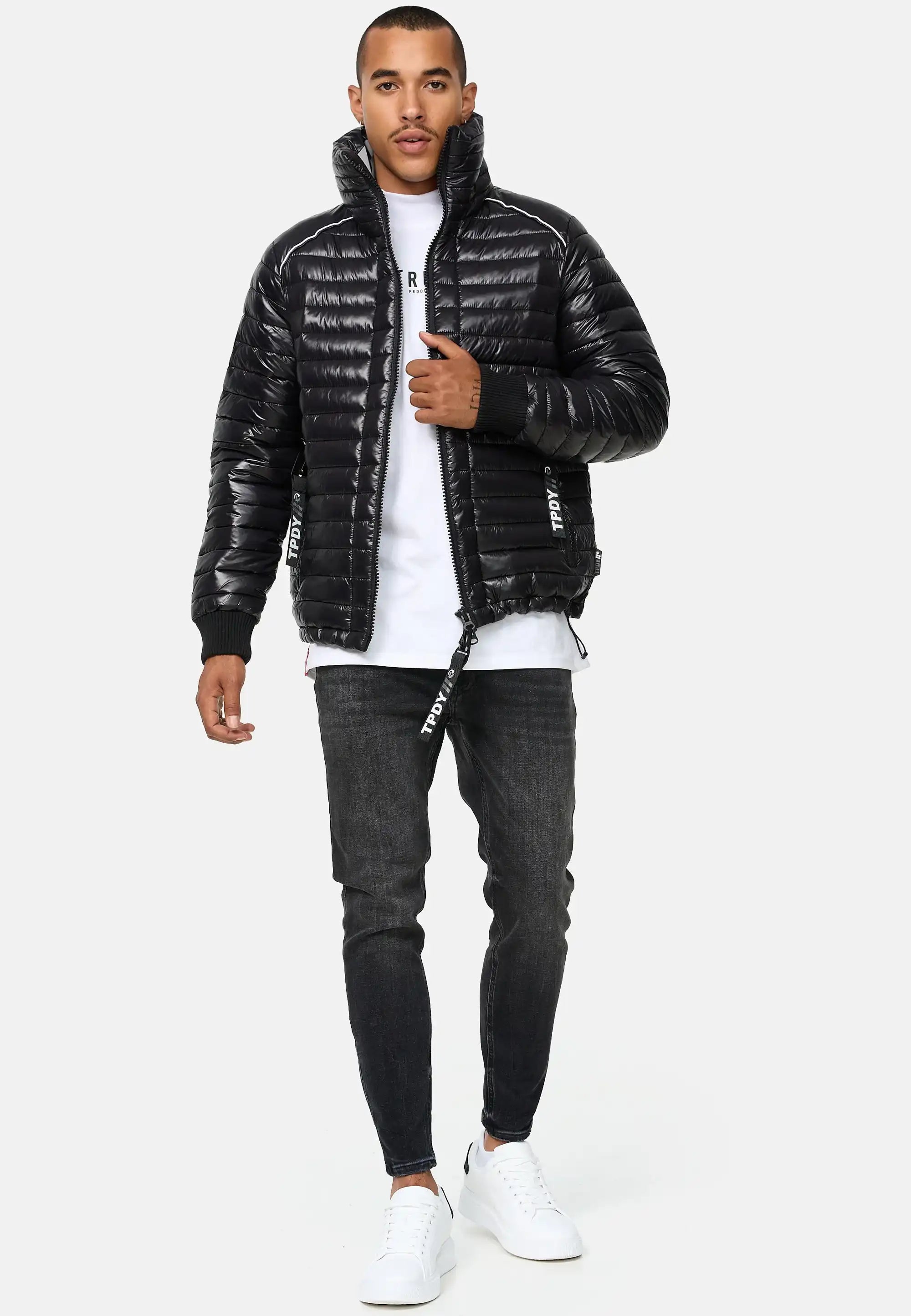 Ein Mann steht mit dem Gesicht nach vorne und trägt die Trueprodigy Corey Steppjacke mit Stehkragen, gepaart mit einem weißen T-Shirt, dunklen Skinny Jeans und weißen Turnschuhen. Eine Hand steckt in seiner Tasche, die andere hält den Reißverschluss der Jacke vor einem hellen Hintergrund.