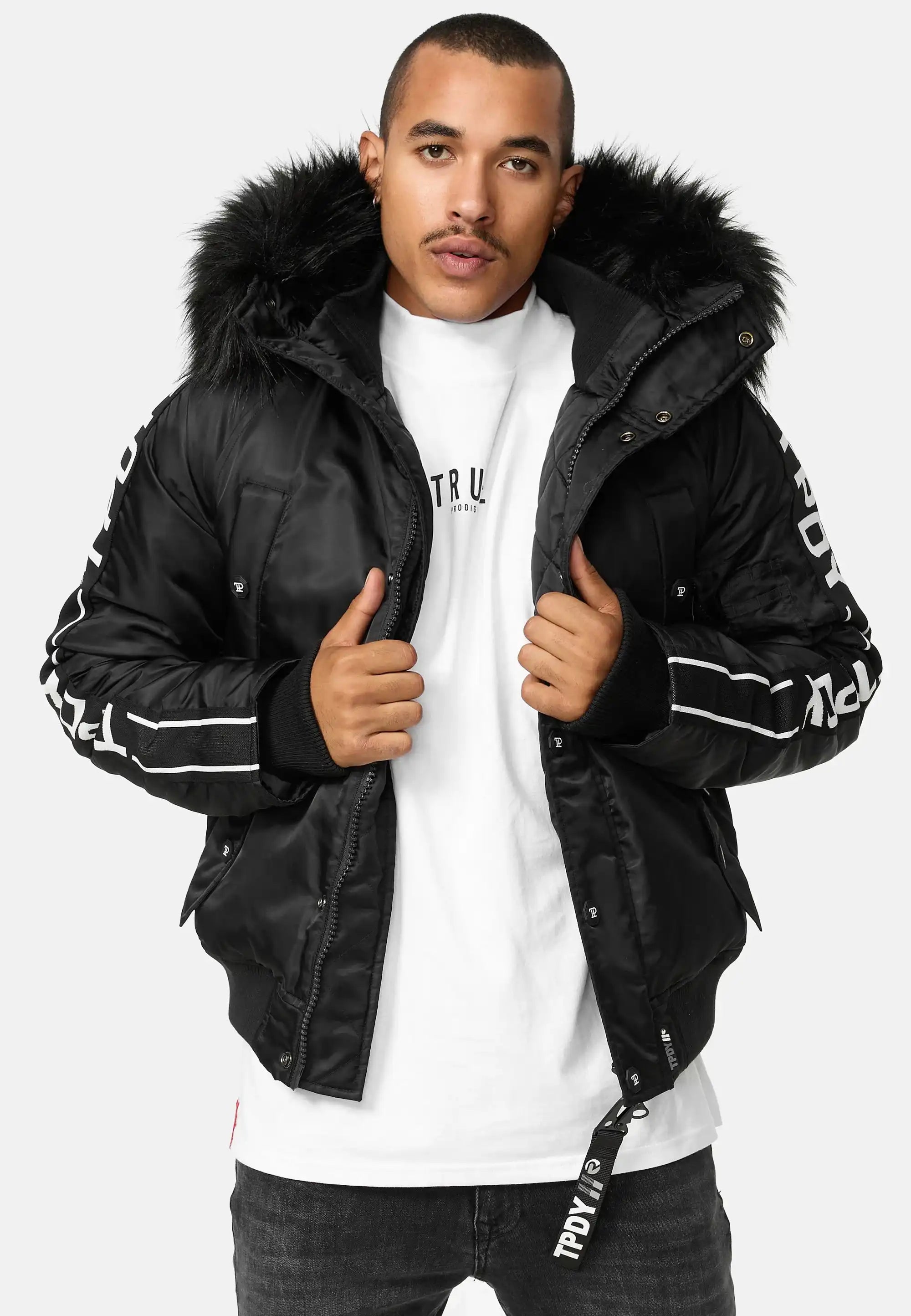 Ein Mann, der die trueprodigy Jonathan Bomberjacke mit Kapuze und Kunstfell, ein weißes T-Shirt und eine dunkle Jeans trägt, steht vor der Kamera und hält seine Jacke offen.