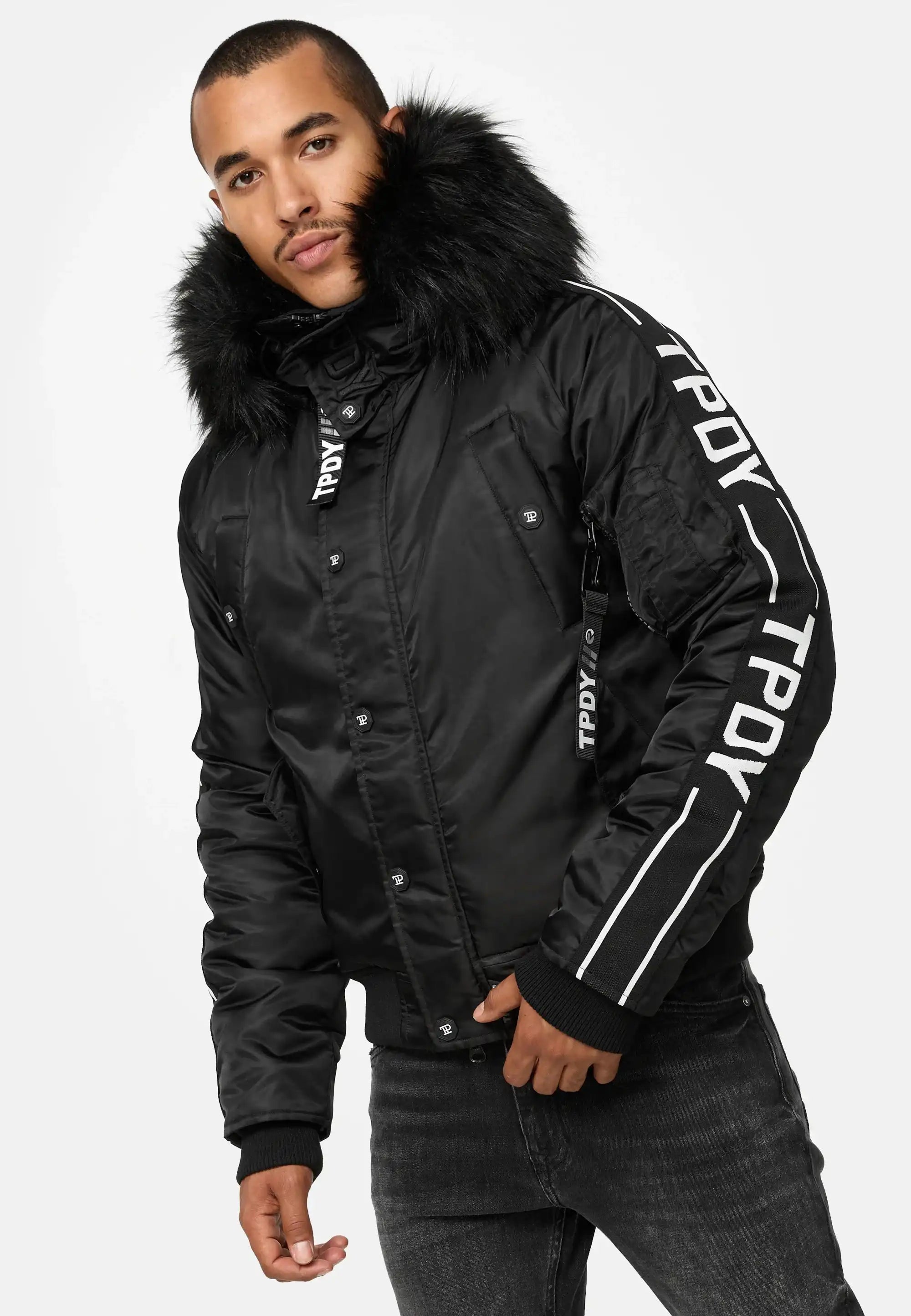 Ein Mann trägt die Jonathan Bomberjacke mit Kapuze und Kunstfell von trueprodigy sowie schwarze Jeans und steht vor einem schlichten weißen Hintergrund.