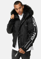 Eine Person, die die Jonathan Bomberjacke mit Kapuze und Kunstfell von trueprodigy mit dem Text TPDY auf den Ärmeln und dem Reißverschluss trägt, über einem weißen Hemd und einer dunklen Jeans, steht vor einem schlichten hellen Hintergrund.