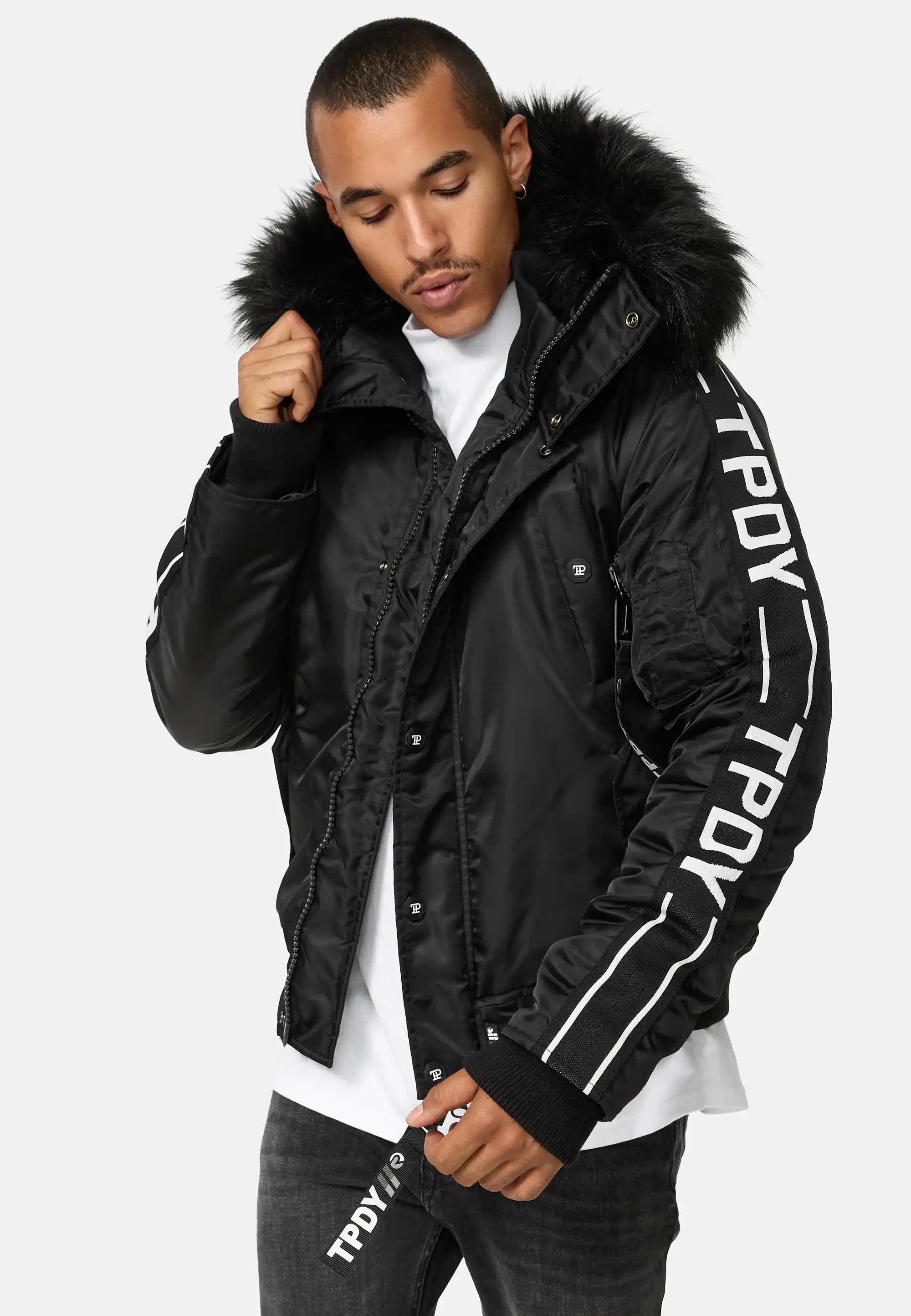 Eine Person, die die Jonathan Bomberjacke mit Kapuze und Kunstfell von trueprodigy mit dem Text TPDY auf den Ärmeln und dem Reißverschluss trägt, über einem weißen Hemd und einer dunklen Jeans, steht vor einem schlichten hellen Hintergrund.