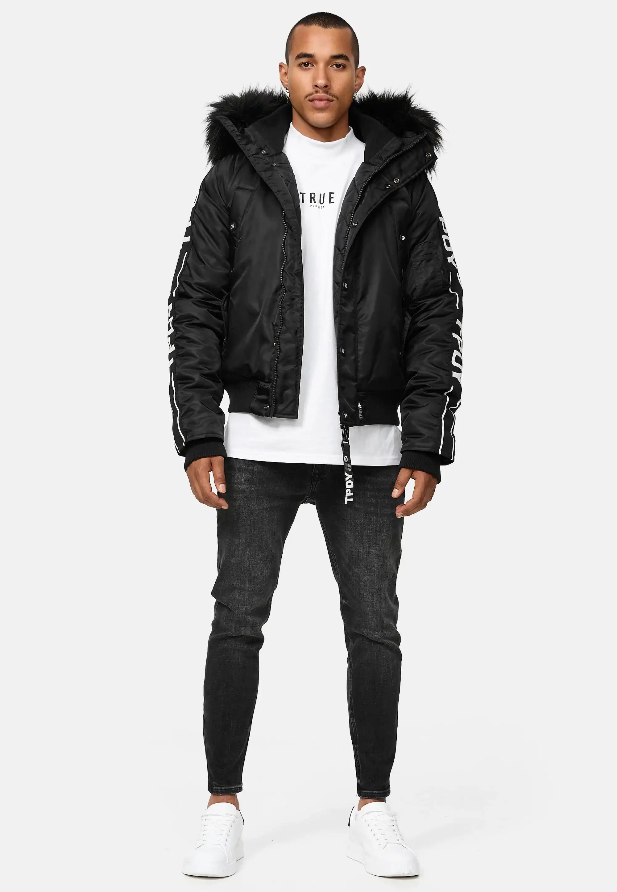 Ein Mann trägt die Jonathan Bomberjacke mit Kapuze und Kunstfell von trueprodigy, dazu ein weißes Shirt, schwarze Skinny Jeans und weiße Sneaker vor schlichtem weißen Hintergrund.