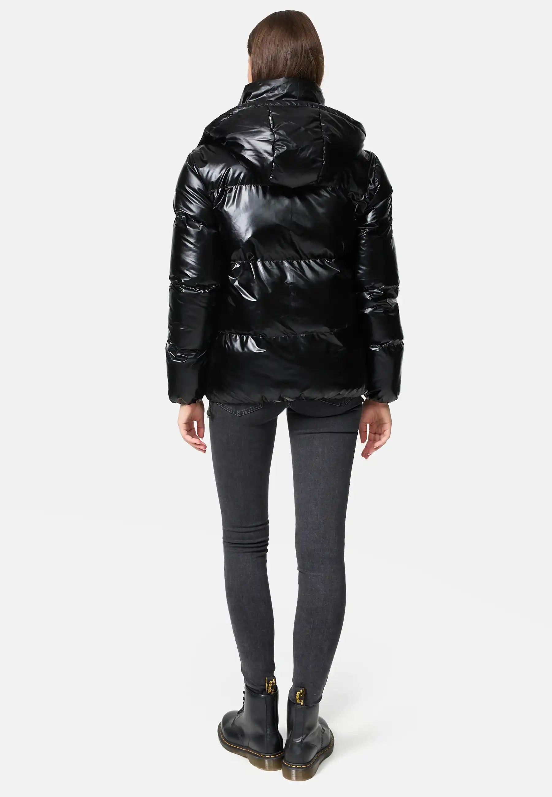 Frau trägt TRUEPRODIGY constance schwarze Steppjacke, Schriftzug „TRUEDPDE“, kombiniert mit schwarzen Jeans und Stiefeln. Jacke glänzend, elegant, wintertauglich, urbaner Stil.