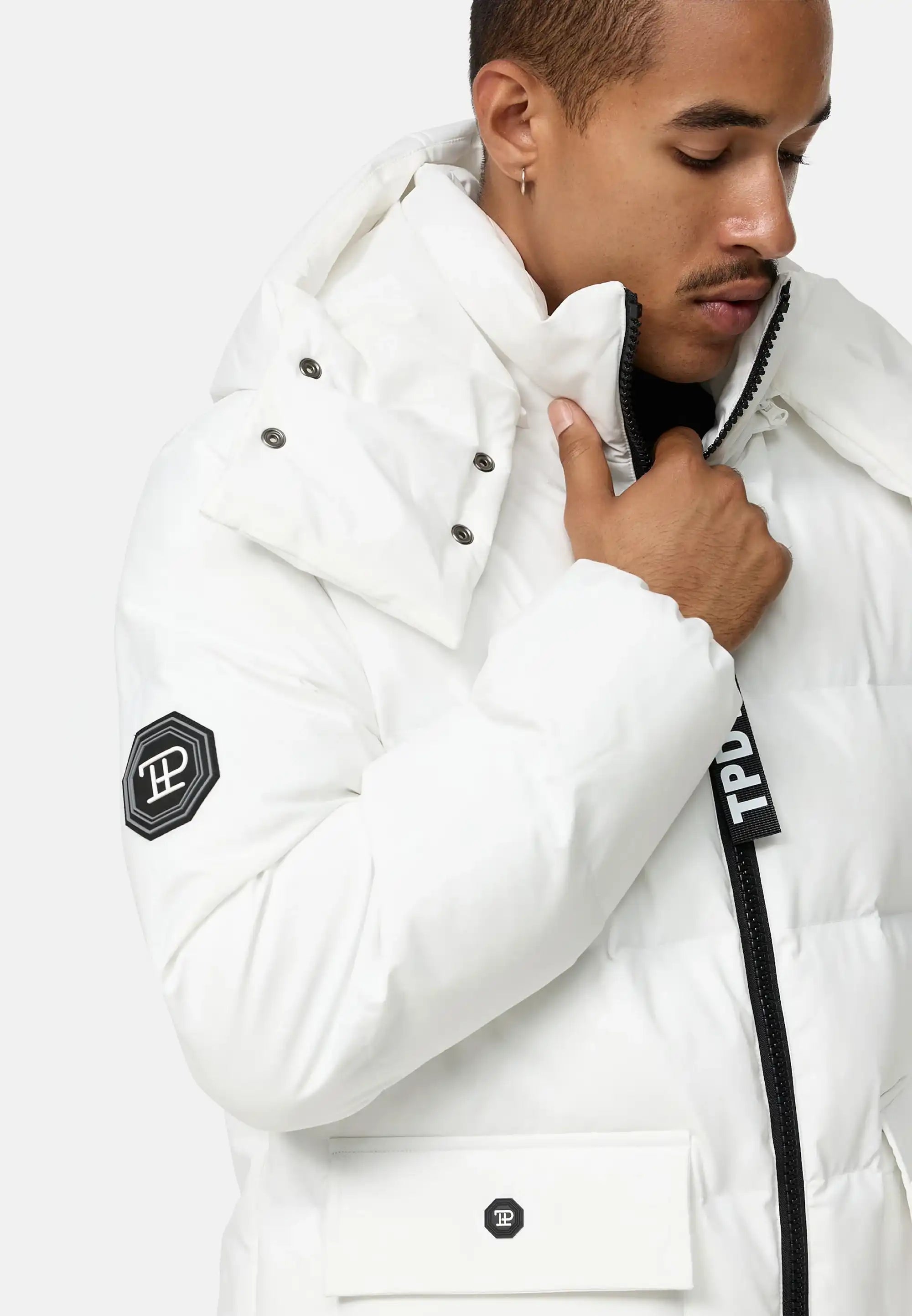 Eine Person in der Trueprodigy Franco Winterjacke mit abnehmbarer Kapuze, weiß mit schwarzen Reißverschlüssen und Logo-Patches an Ärmel und Tasche, steht vor hellem Hintergrund, blickt nach unten und hält den Kragen.