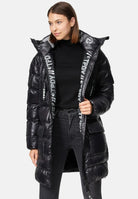 Eine Frau trägt die trueprodigy David F Glänzende Steppjacke mit Kapuze mit hohem Kragen über einem schwarzen Rollkragen und dunklen Jeans. Das Innenfutter der Jacke ist mit weißer Schrift versehen, während sie leicht zur Seite gewandt steht.