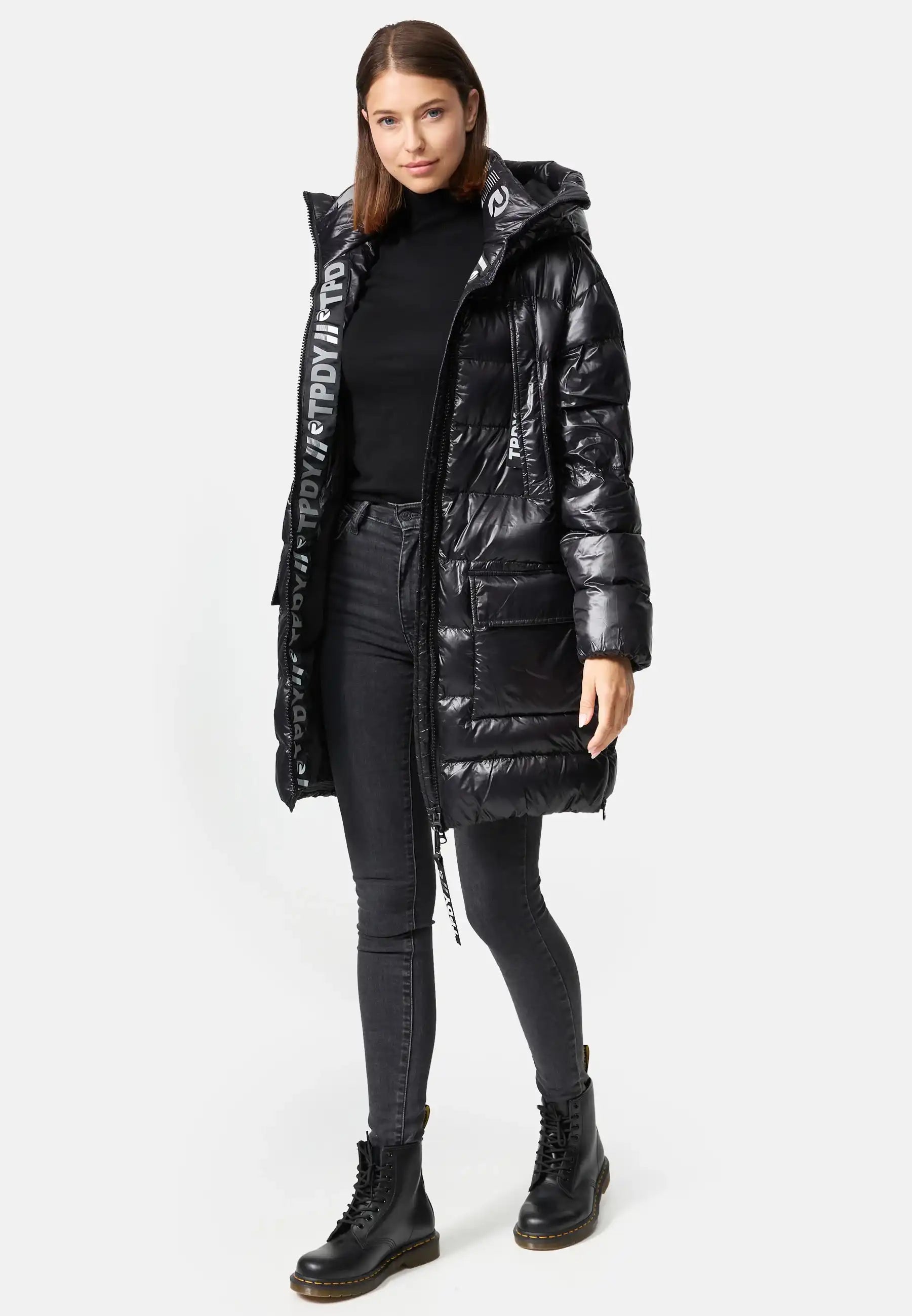 Eine Frau blickt nach vorne und trägt die trueprodigy David F Glänzende Steppjacke mit Kapuze mit großen Taschen auf der Vorderseite und Text auf der Innenkapuze. Dazu trägt sie einen schwarzen Rollkragenpullover, graue Skinny Jeans und schwarze Schnürstiefel.