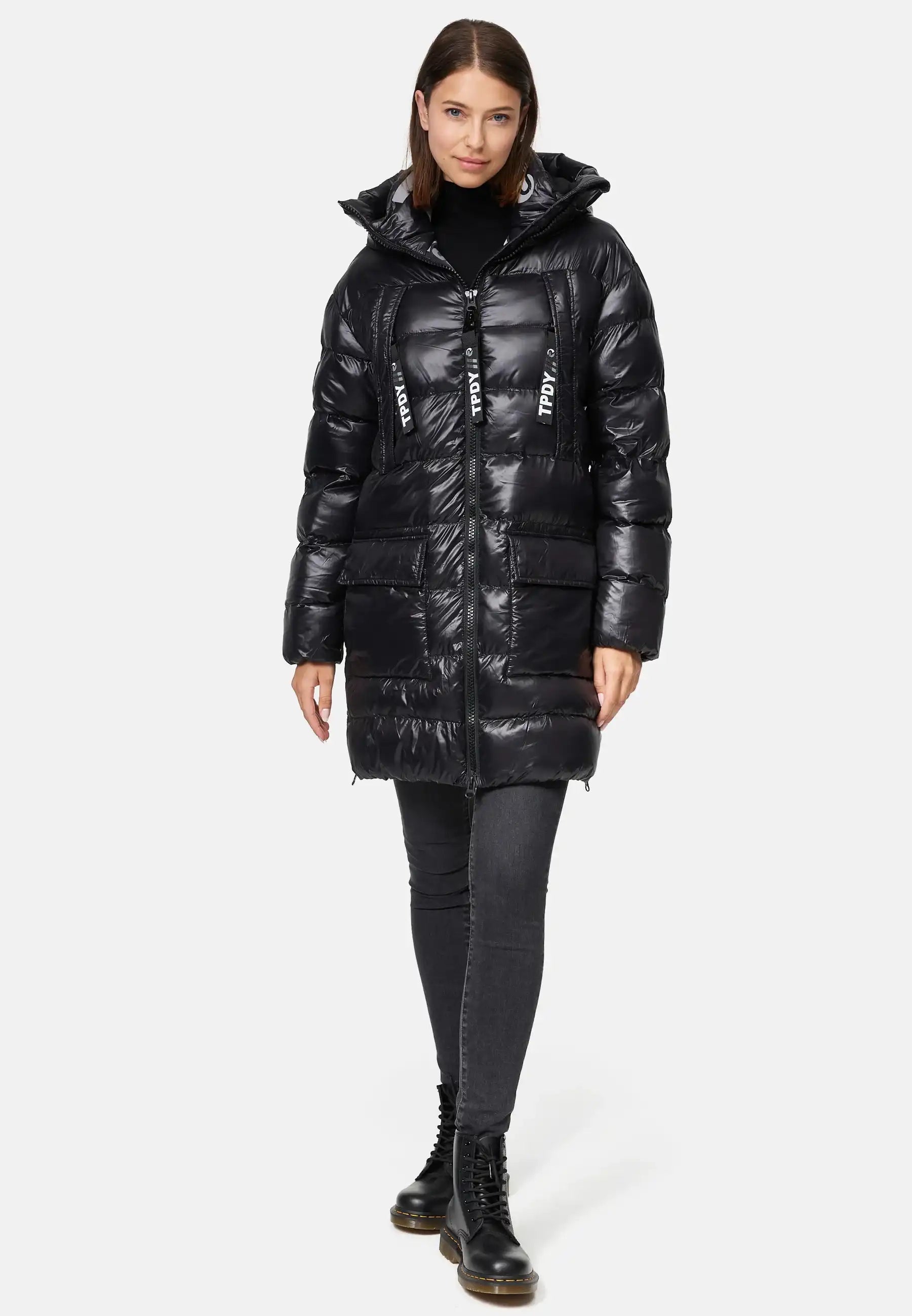 Eine Frau blickt nach vorne in der trueprodigy David F Glänzende Steppjacke mit Kapuze, einem glänzenden schwarzen Puff mit Kapuze, großen Reißverschlüssen mit weißem Text, gepaart mit schwarzen Jeans und Schnürstiefeln. Ihr Haar ist glatt und schulterlang.