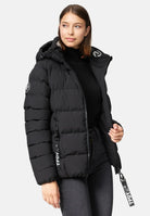 Eine Frau trägt die schwarze Murphy F Matte Steppjacke mit Kapuze von trueprodigy, wasserabweisend mit sichtbaren TPDY-Zippern und rundem Ärmel-Aufnäher, dazu einen schwarzen Rollkragen und dunkle Jeans vor hellem Hintergrund.