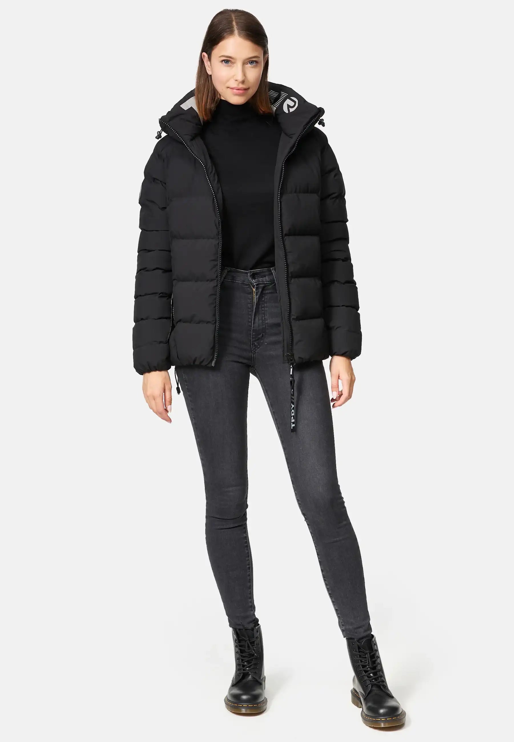 Eine Frau trägt die Murphy F Matte Steppjacke mit Kapuze von trueprodigy, kombiniert mit schwarzem Rollkragen, dunklen Skinny-Jeans und schwarzen Schnürstiefeln. Sie hat glattes, schulterlanges braunes Haar und einen neutralen Ausdruck.