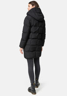 Eine Frau steht mit dem Gesicht zur Wand und trägt eine Trueprodigy Willo F Lange Pufferjacke mit Kapuze, dunkle Skinny-Jeans und schwarze Ankle Boots vor einem schlichten weißen Hintergrund.