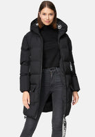 Eine Frau mit glattem braunem Haar blickt nach vorne und trägt die trueprodigy Willo F Lange Pufferjacke mit Kapuze, einen langen schwarzen Mantel mit Reißverschlüssen, Taschen, weißen Textdetails am Ärmel und Träger, über einem schwarzen Rollkragenpullover und dunklen Jeans.