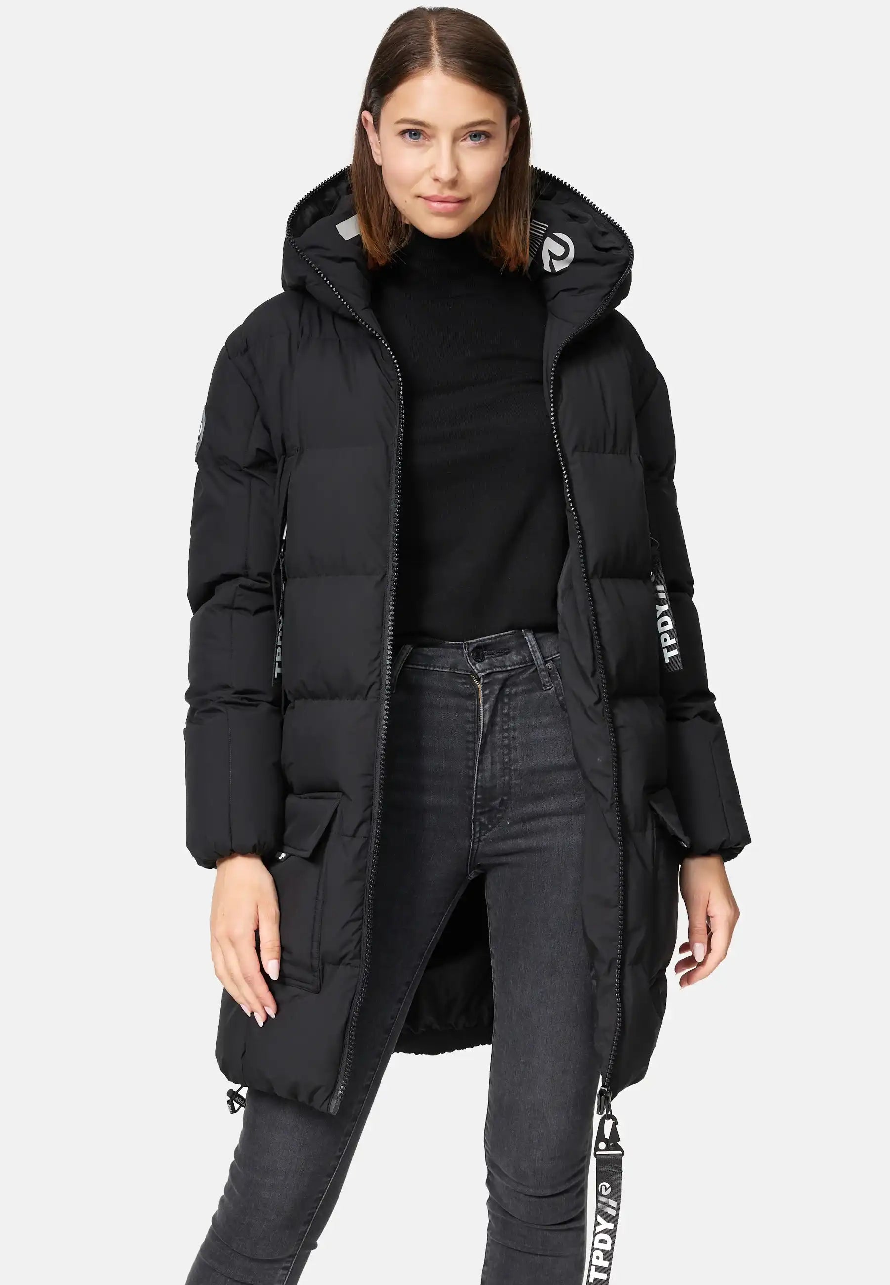 Eine Frau mit glattem braunem Haar blickt nach vorne und trägt die trueprodigy Willo F Lange Pufferjacke mit Kapuze, einen langen schwarzen Mantel mit Reißverschlüssen, Taschen, weißen Textdetails am Ärmel und Träger, über einem schwarzen Rollkragenpullover und dunklen Jeans.