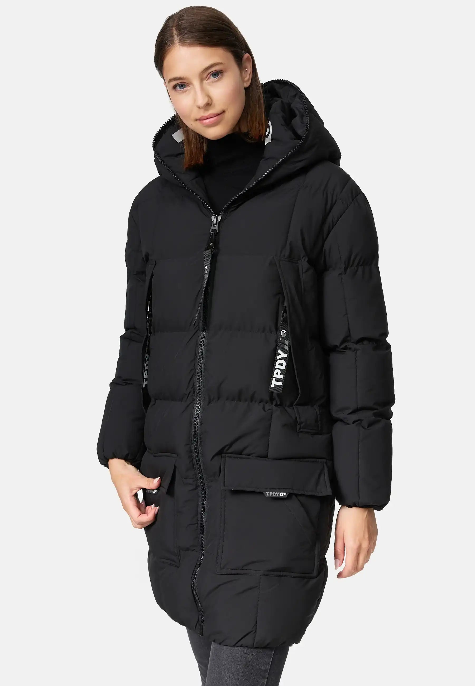 Eine Person steht in einer schwarzen Trueprodigy Willo F Lange Pufferjacke mit Kapuze, die eine Kapuze, einen Frontreißverschluss, Brusttaschen mit Reißverschluss, einen hohen Kragen und zwei große Fronttaschen aufweist. Eine Hand ruht in einer Tasche. Der Hintergrund ist einfarbig weiß.