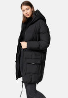 Eine Frau mit glattem braunem Haar trägt die trueprodigy Willo F Lange Pufferjacke mit Kapuze in Schwarz über einer dunklen Jeans, wobei sie leicht zur Seite schaut und den Reißverschluss festhält. Der Hintergrund ist einfarbig hellgrau.