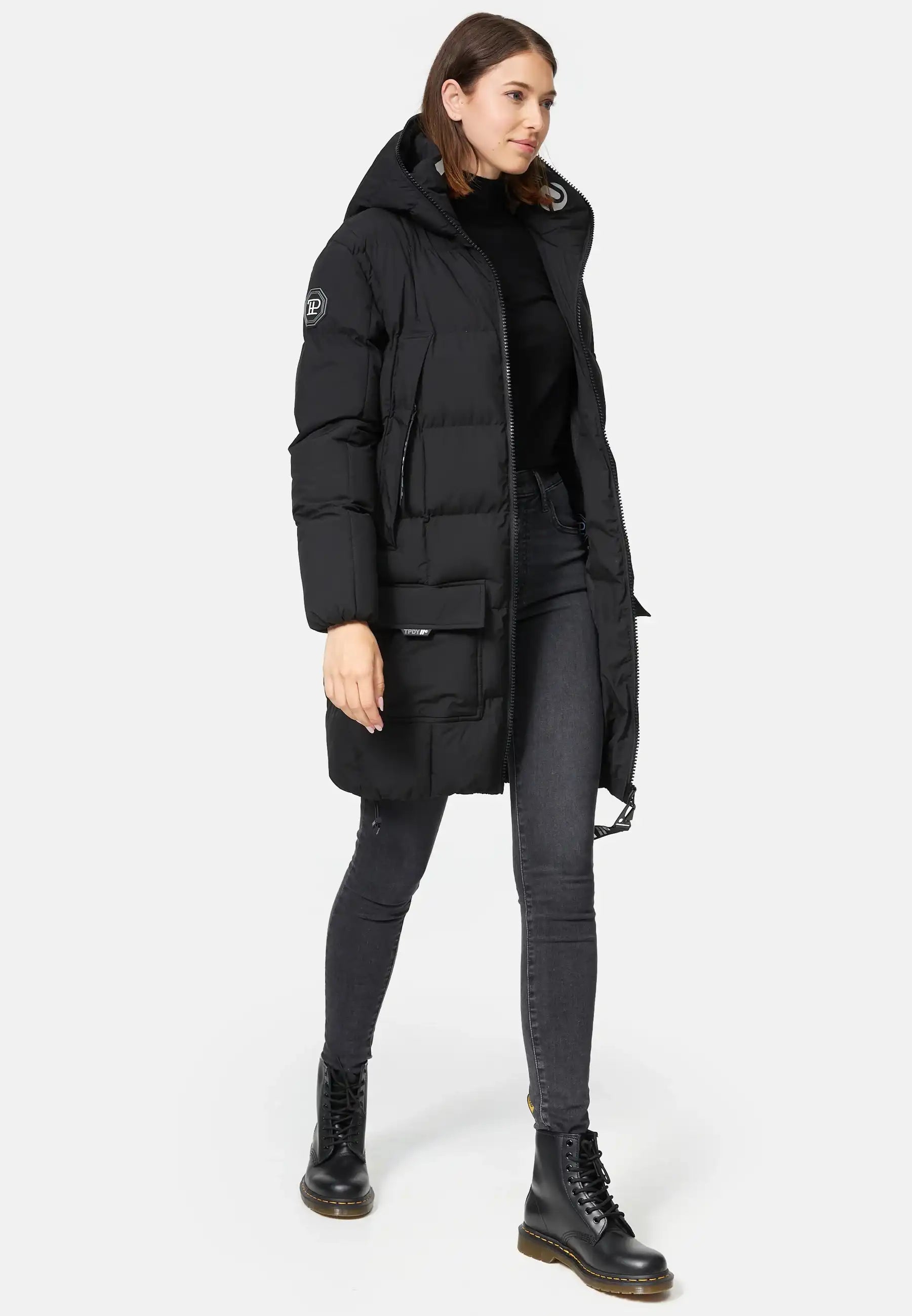 Eine Person, die die trueprodigy Willo F Lange Pufferjacke mit Kapuze in Schwarz trägt, gestylt mit einem schwarzen Rollkragenpullover, dunklen Skinny Jeans und schwarzen Schnürstiefeln, steht vor einem schlichten weißen Hintergrund.
