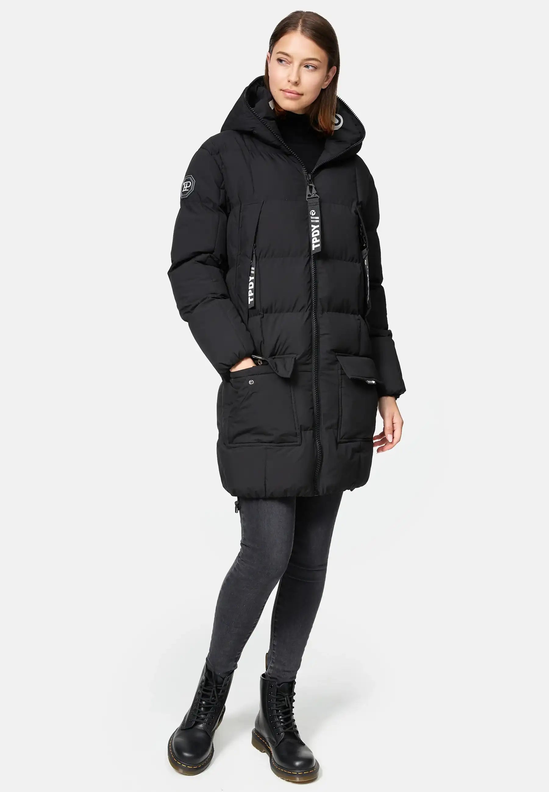 Eine Frau trägt die Willo F Lange Pufferjacke mit Kapuze von trueprodigy zu dunklen Skinny-Jeans und schwarzen Schnürstiefeln. Die Jacke ist wasserabweisend, hat große Taschen sowie sichtbare Reißverschlüsse. Der Hintergrund ist weiß.
