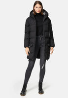 Eine Frau steht vor einem schlichten Hintergrund und trägt die trueprodigy Willo F Lange Pufferjacke mit Kapuze, gepaart mit einem schwarzen Rollkragenpullover, dunklen Skinny Jeans und schwarzen Schnürstiefeln. Ihre Hände liegen entspannt an ihren Seiten.