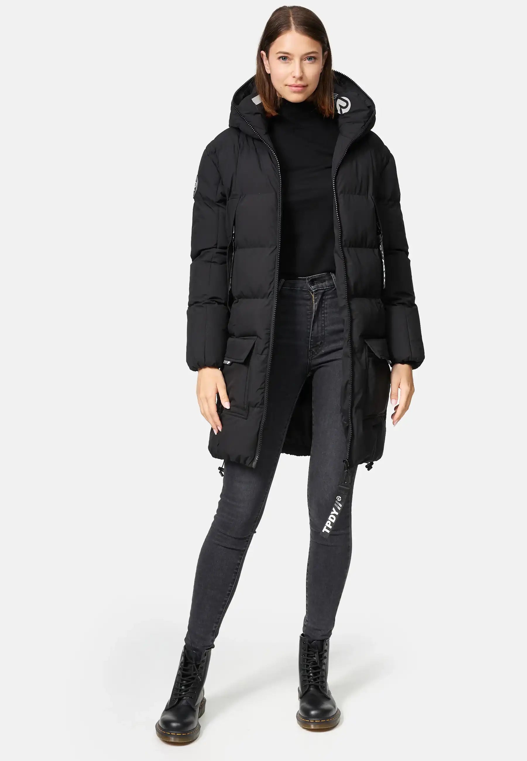 Eine Frau steht vor einem schlichten Hintergrund und trägt die trueprodigy Willo F Lange Pufferjacke mit Kapuze, gepaart mit einem schwarzen Rollkragenpullover, dunklen Skinny Jeans und schwarzen Schnürstiefeln. Ihre Hände liegen entspannt an ihren Seiten.