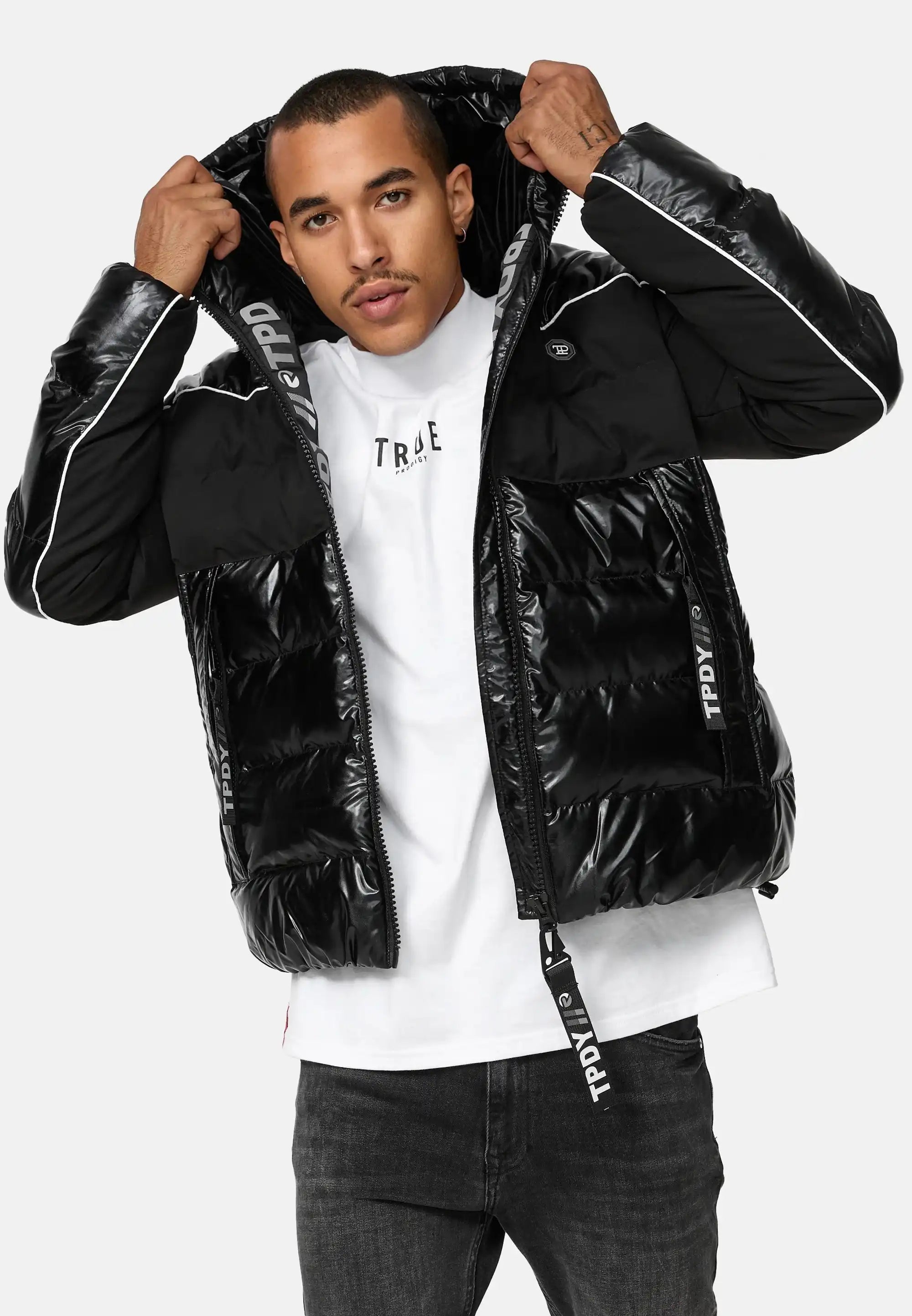 Ein Mann steht mit dem Gesicht nach vorne und trägt eine glänzende schwarze trueprodigy Clark Winterjacke mit Kapuze und Logo mit weißen Paspeln und gebrandeten Reißverschlüssen über einem weißen T-Shirt. Er hält die Kapuze der reflektierenden Jacke mit beiden Händen fest.