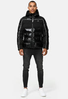 Ein Mann blickt nach vorne und trägt die glänzende schwarze trueprodigy Clark Winterjacke mit Kapuze und Logo mit weißen Paspeln, dunkle Skinny Jeans und weiße Turnschuhe vor einem schlichten hellgrauen Hintergrund.