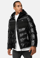 Ein Mann in der glänzenden Clark Winterjacke mit Kapuze und Logo von trueprodigy mit weißen Paspeln und einer Kapuze über einem weißen Hemd und einer dunklen Jeans steht leicht seitlich vor einem unifarbenen Hintergrund.