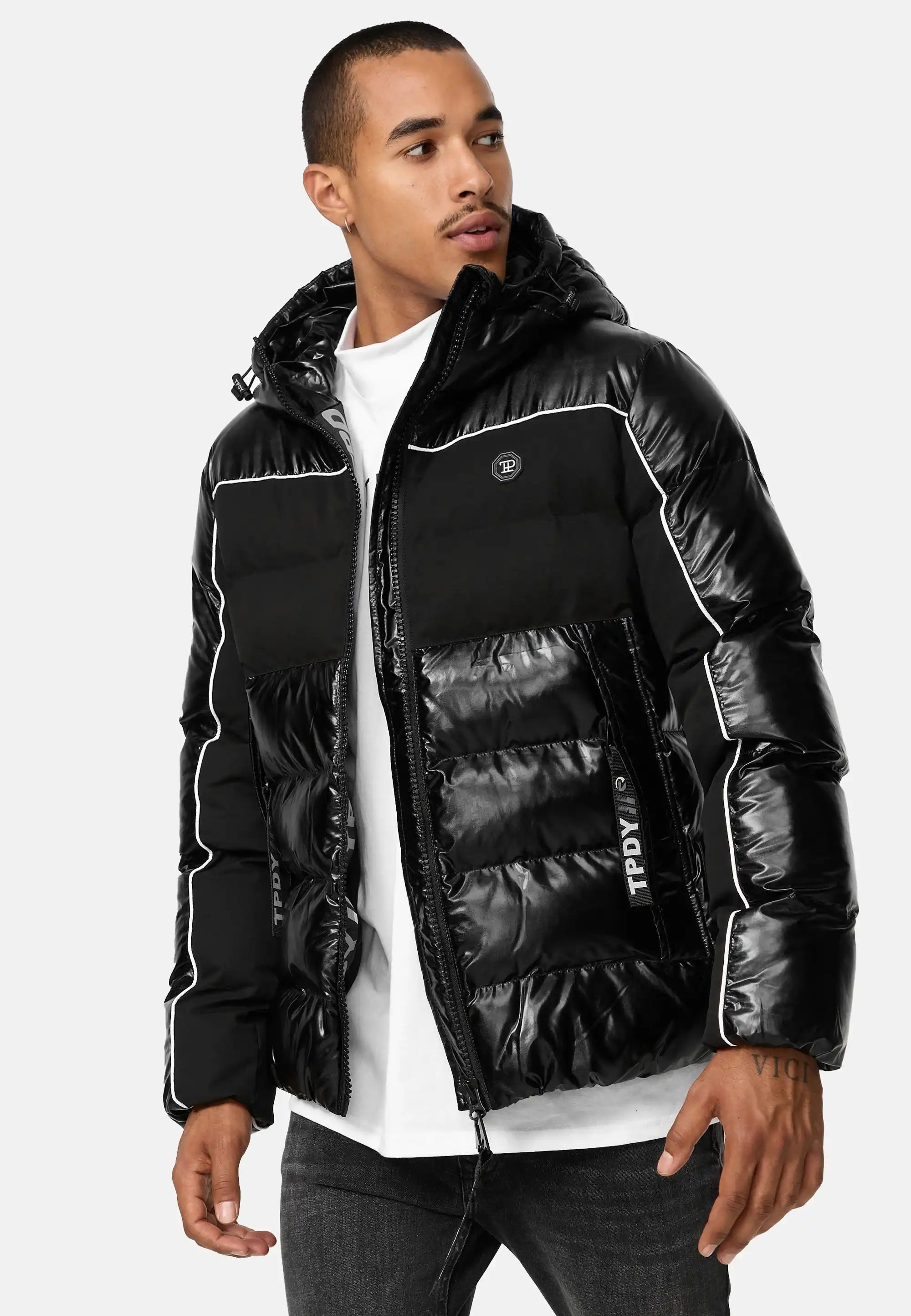 Ein Mann in der glänzenden Clark Winterjacke mit Kapuze und Logo von trueprodigy mit weißen Paspeln und einer Kapuze über einem weißen Hemd und einer dunklen Jeans steht leicht seitlich vor einem unifarbenen Hintergrund.
