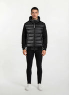 A man steht frontal vor weißem Hintergrund, trägt eine gesteppte Herren Übergangsjacke für urbanen City-Style von trueprodigy mit Kapuze, dazu schwarze Hosen und weiße Sneaker. Seine Arme hängen locker an den Seiten.