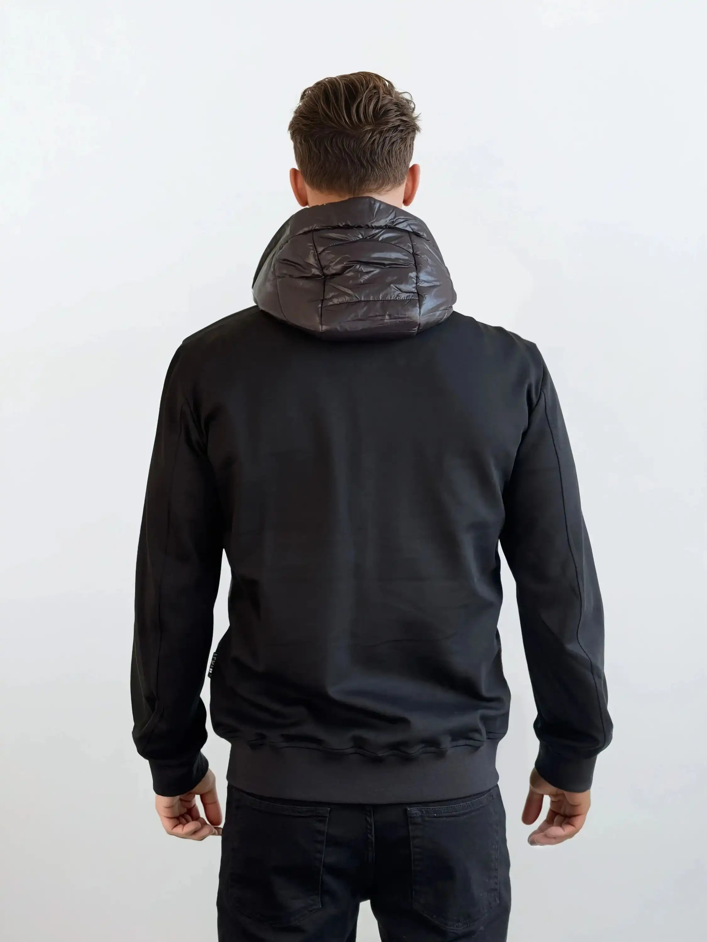 Ein Mann steht mit dem Rücken zur Kamera, bekleidet mit einer trueprodigy Gesteppte Herren Übergangsjacke für urbanen City-Style und einer schwarzen Hose. Der Hintergrund ist einfarbig weiß.