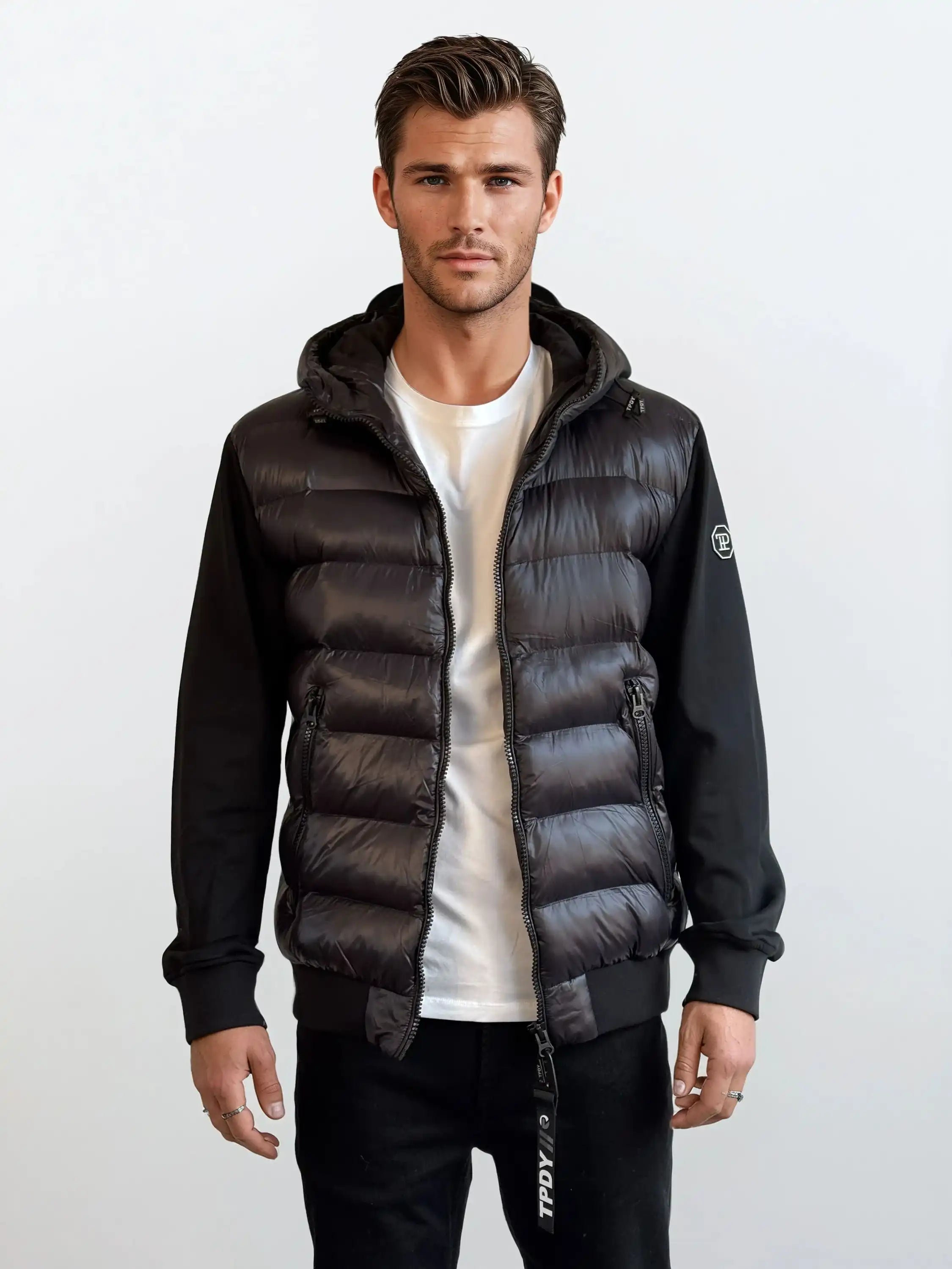 Ein Mann trägt eine schwarze Gesteppte Herren Übergangsjacke für urbanen City-Style von trueprodigy mit weißem T-Shirt und schwarzen Hosen. Der Hintergrund ist schlicht und hell gehalten.