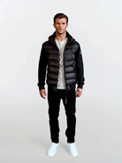 Ein Mann blickt nach vorne in einer trueprodigy Gesteppte Herren Übergangsjacke für urbanen City-Style über einem weißen T-Shirt, einer schwarzen Hose und weißen Sneakers. Der Hintergrund ist schlicht und hell.