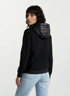 Eine Person mit mittellangen dunklen Haaren trägt eine trueprodigy Oversized Damen Bomberjacke im urbanen Materialmix-Design und eine hellblaue Jeans. Sie steht mit dem Rücken zur Kamera vor einem schlichten hellen Hintergrund.