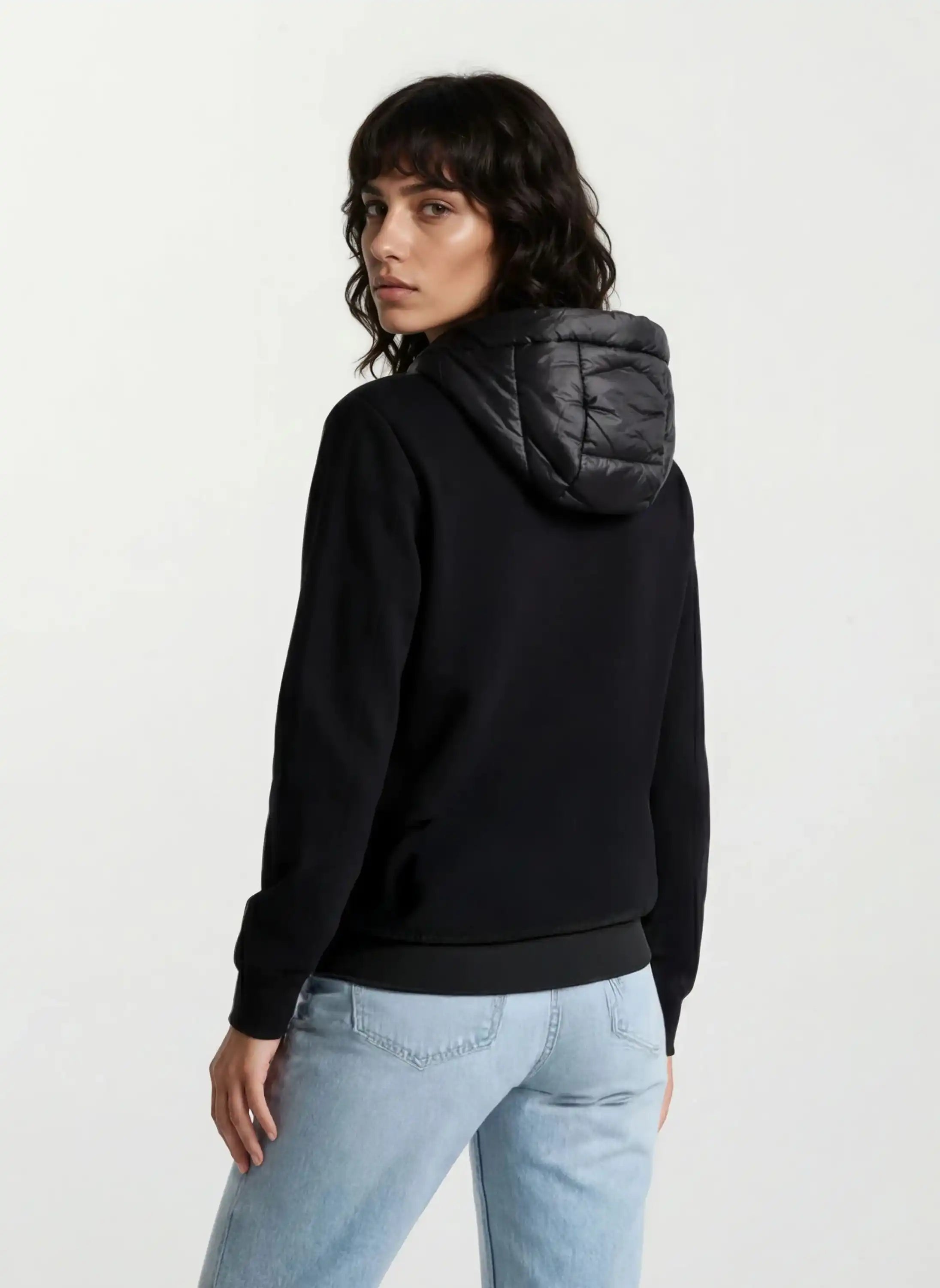 Eine Person mit mittellangen dunklen Haaren trägt eine trueprodigy Oversized Damen Bomberjacke im urbanen Materialmix-Design und eine hellblaue Jeans. Sie steht mit dem Rücken zur Kamera vor einem schlichten hellen Hintergrund.