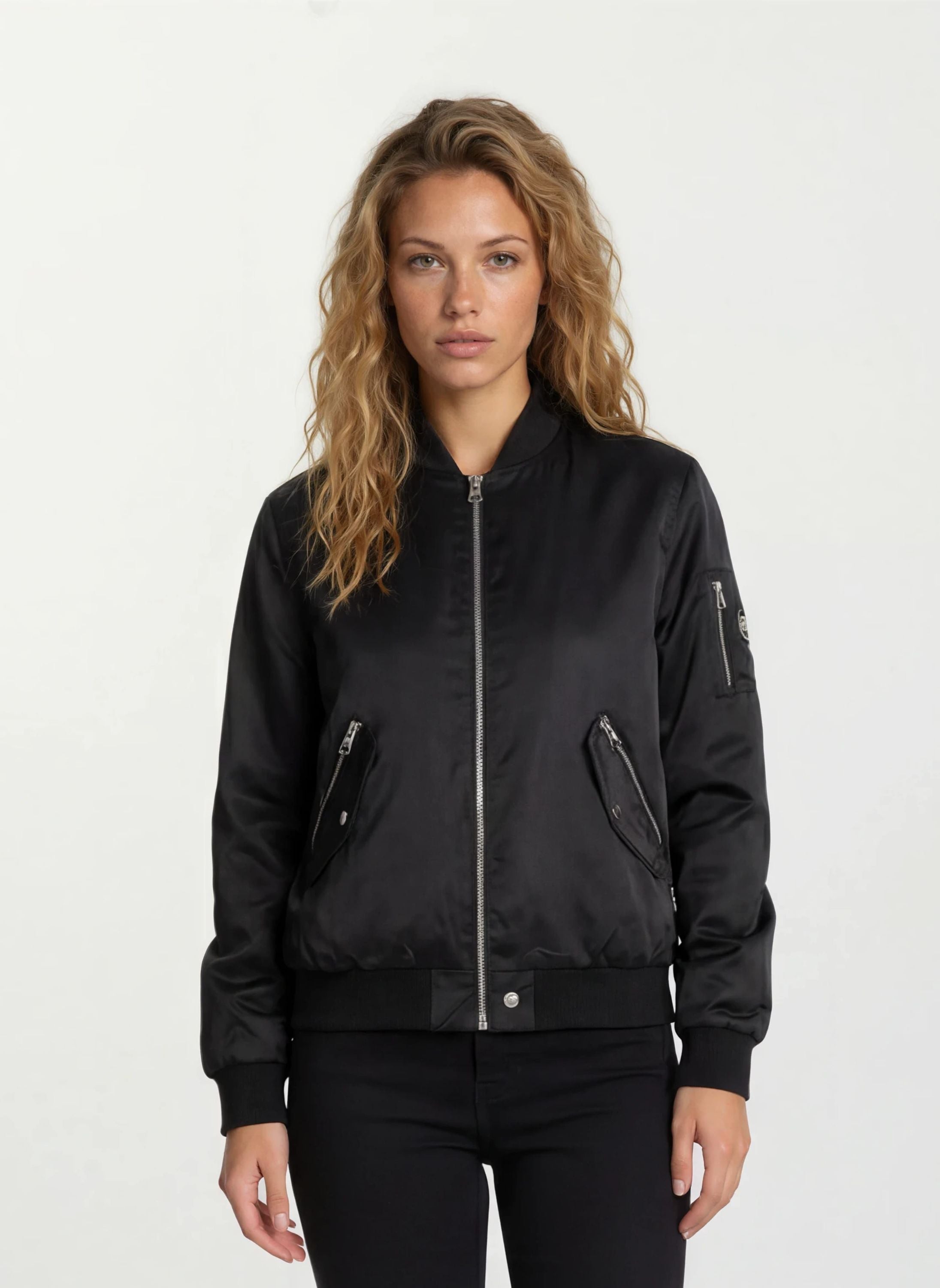 Eine Frau mit gewelltem, hellbraunem Haar steht vor einem unifarbenen Hintergrund, trägt die trueprodigy Elegante Damen Bomber Jacke mit glänzender Oberfläche und eine schwarze Hose und zeigt einen modernen, urbanen Stil, während sie direkt in die Kamera schaut.