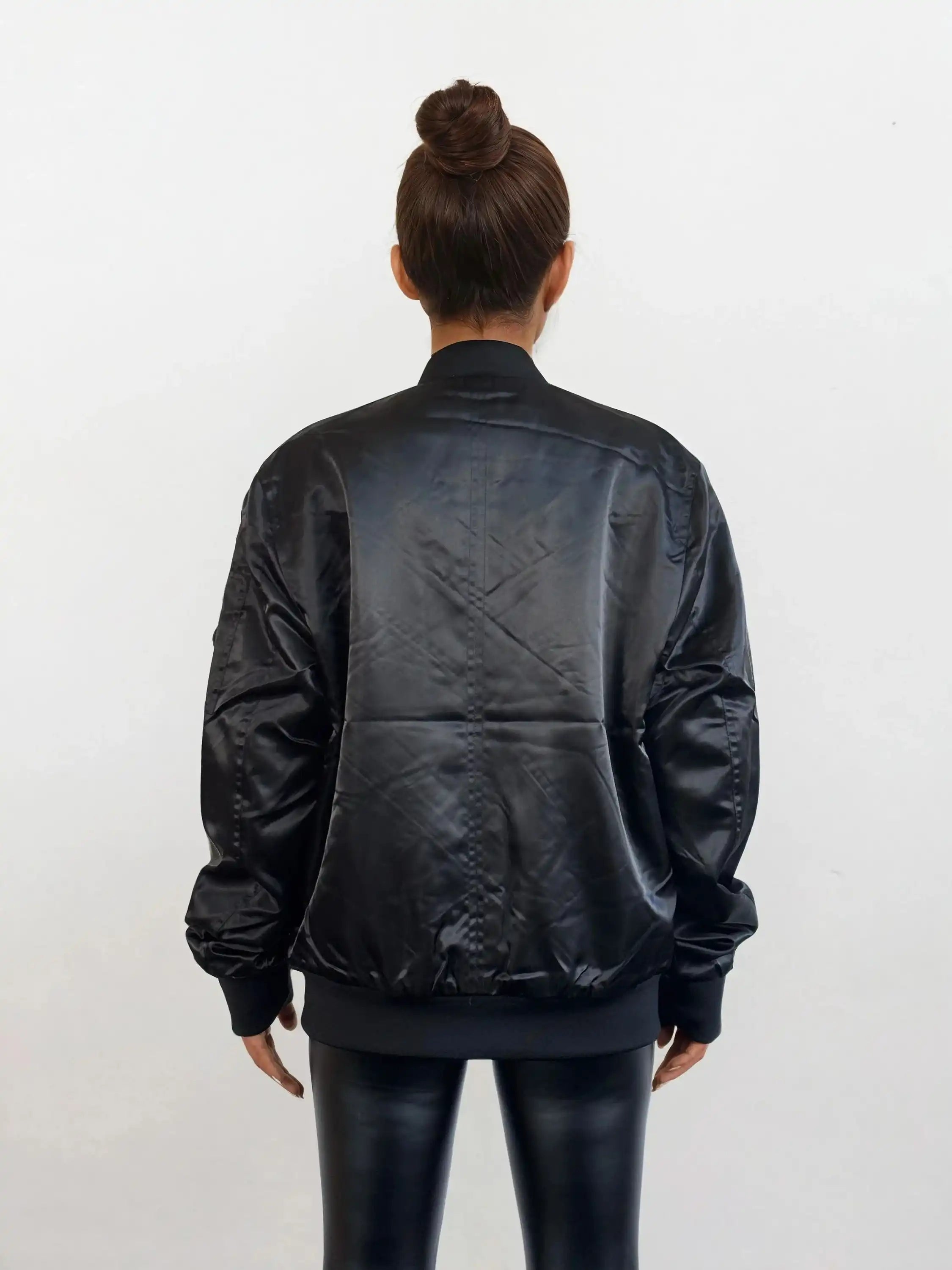 Eine Frau mit gepflegtem Dutt, von hinten gesehen vor einem weißen Hintergrund, trägt glänzende schwarze Leggings und die Elegante Damen Bomber Jacke mit glänzender Oberfläche von trueprodigy.