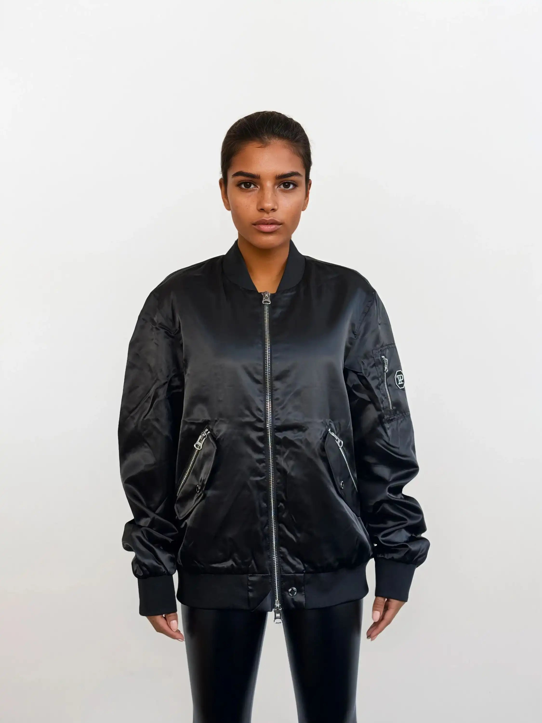 Eine Frau steht mit dem Gesicht nach vorne vor einem schlichten weißen Hintergrund und trägt eine trueprodigy Elegante Damen Bomberjacke mit glänzender Oberfläche, gepaart mit einer glänzenden schwarzen Hose.