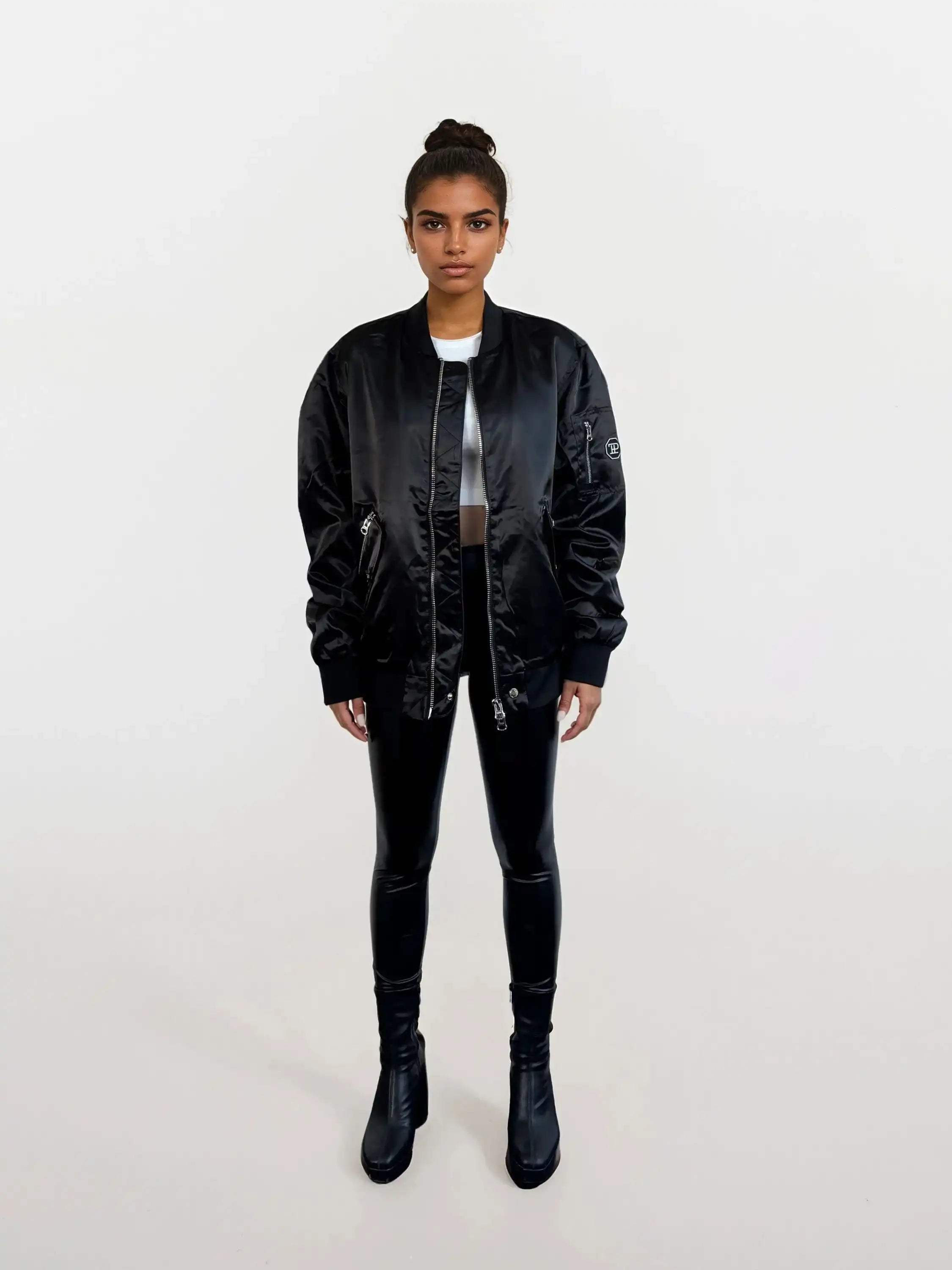 Eine Person steht mit dem Gesicht nach vorne vor einem weißen Hintergrund, trägt eine schwarze trueprodigy Elegante Damen Bomberjacke mit glänzender Oberfläche, schwarz glänzende Leggings, ein weißes Oberteil, schwarze Stiefel und hat die Haare zu einem Dutt gebunden.