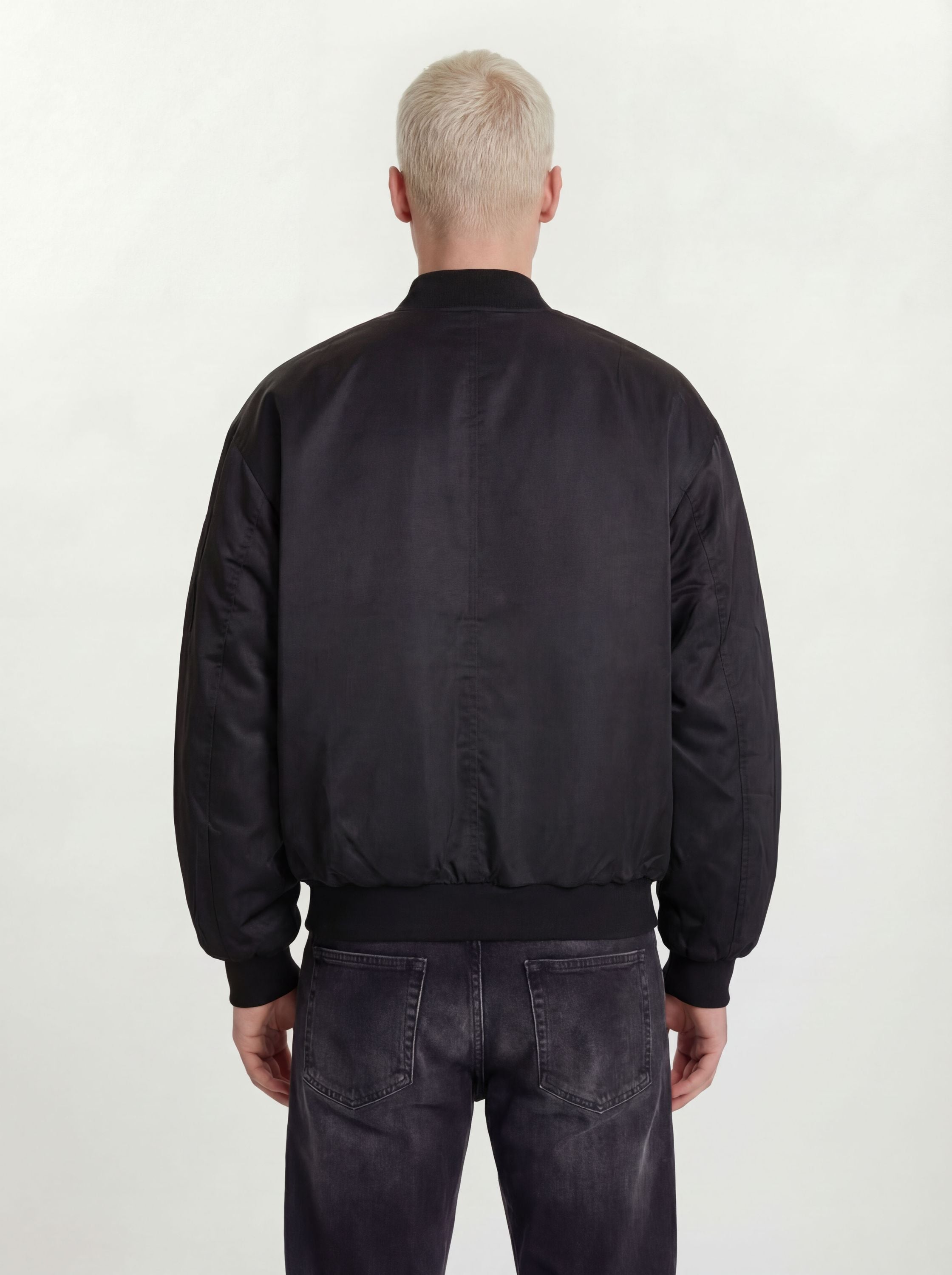 Eine Person mit kurzen, hellblonden Haaren ist von hinten zu sehen. Sie trägt eine trueprodigy Klassische Herren Bomberjacke mit glänzender Oberfläche und dunkel gewaschene Jeans, die vor einem schlichten, hellen Hintergrund für einen urbanen Style-Look stehen.