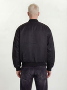 Eine Person mit kurzen, hellblonden Haaren ist von hinten zu sehen. Sie trägt eine trueprodigy Klassische Herren Bomberjacke mit glänzender Oberfläche und dunkel gewaschene Jeans, die vor einem schlichten, hellen Hintergrund für einen urbanen Style-Look stehen.