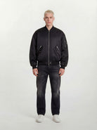 Eine Person mit kurzen blonden Haaren trägt eine trueprodigy Klassische Herren Bomberjacke mit glänzender Oberfläche, dunkle Jeans und weiße Turnschuhe und steht mit dem Gesicht nach vorne vor einem schlichten hellen Hintergrund.