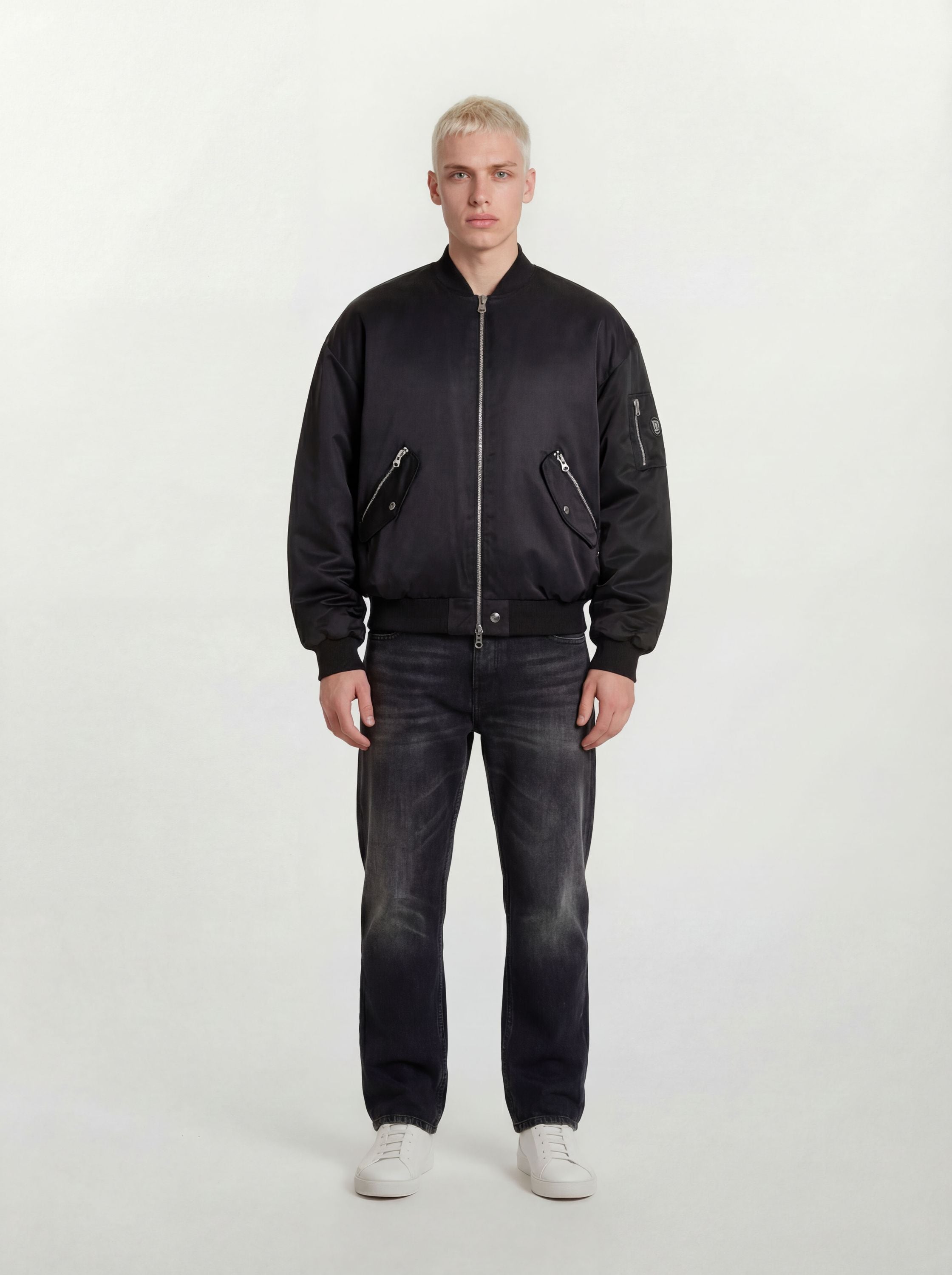 Eine Person mit kurzen blonden Haaren trägt eine trueprodigy Klassische Herren Bomberjacke mit glänzender Oberfläche, dunkle Jeans und weiße Turnschuhe und steht mit dem Gesicht nach vorne vor einem schlichten hellen Hintergrund.