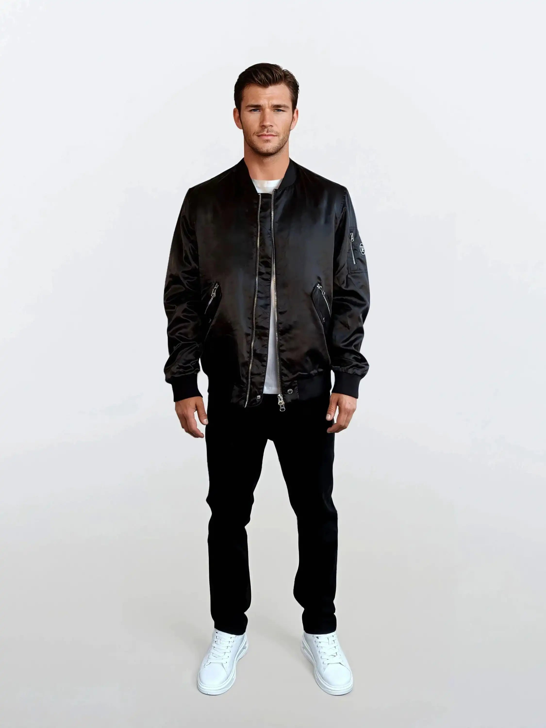 Ein Mann blickt vor einem weißen Hintergrund nach vorne und trägt einen urbanen Style mit einer trueprodigy Klassische Herren Bomber Jacke mit glänzender Oberfläche, einem weißen T-Shirt, einer schwarzen Hose und weißen Turnschuhen. Seine Arme sind entspannt an seinen Seiten.