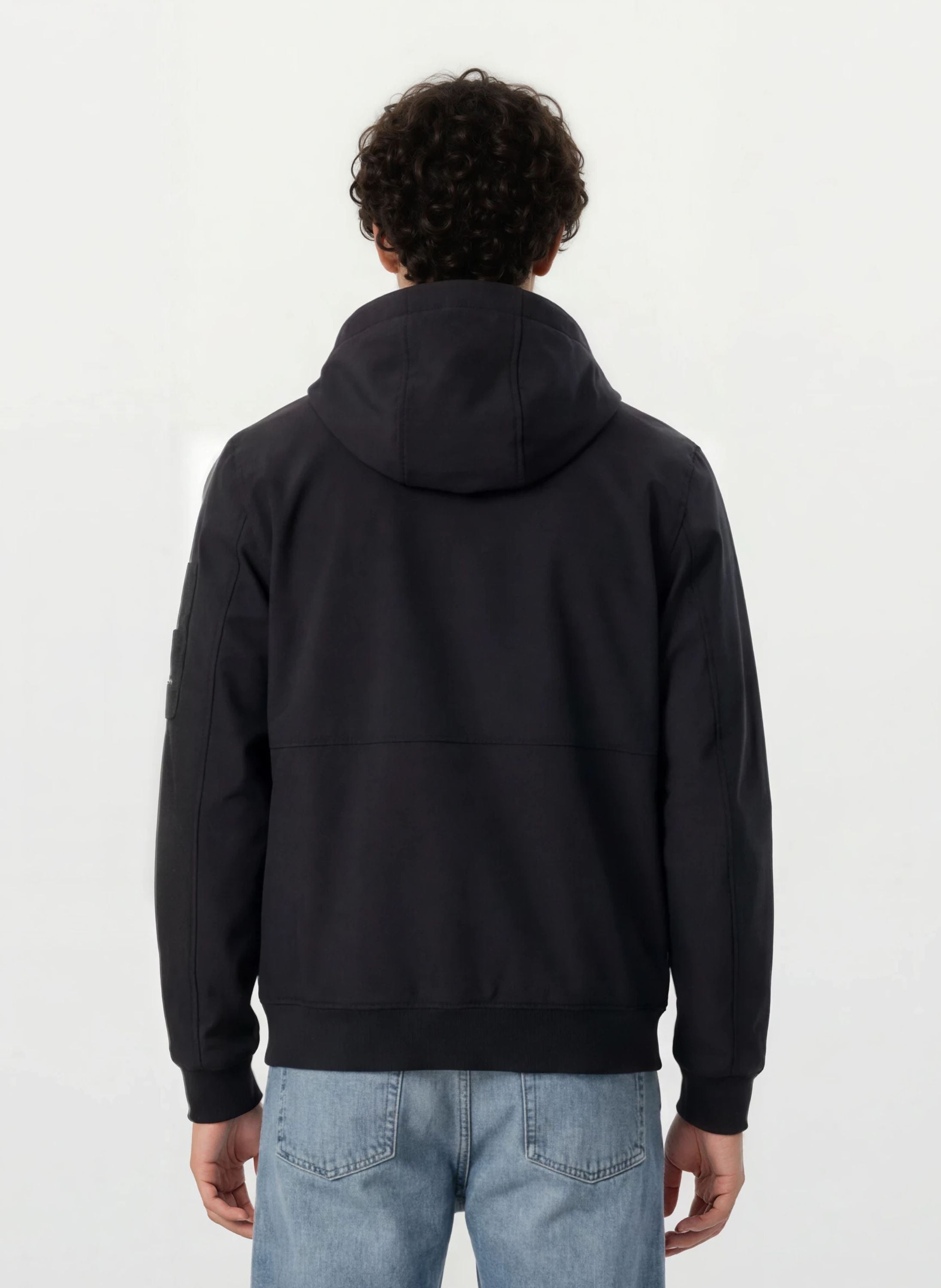 Eine Person mit lockigem Haar wird von hinten gezeigt, die eine Trueprodigy Leichte Herrenjacke mit Brust- und Ärmelbadge und hellblaue Jeans trägt und vor einem schlichten, hellen Hintergrund steht.