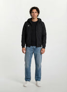 Ein Mann steht mit dem Gesicht nach vorne vor einem schlichten weißen Hintergrund. Er trägt eine Trueprodigy Leichte Herrenjacke mit Brust- und Ärmelbadge über einem schwarzen T-Shirt, blaue Jeans und weiße Turnschuhe.