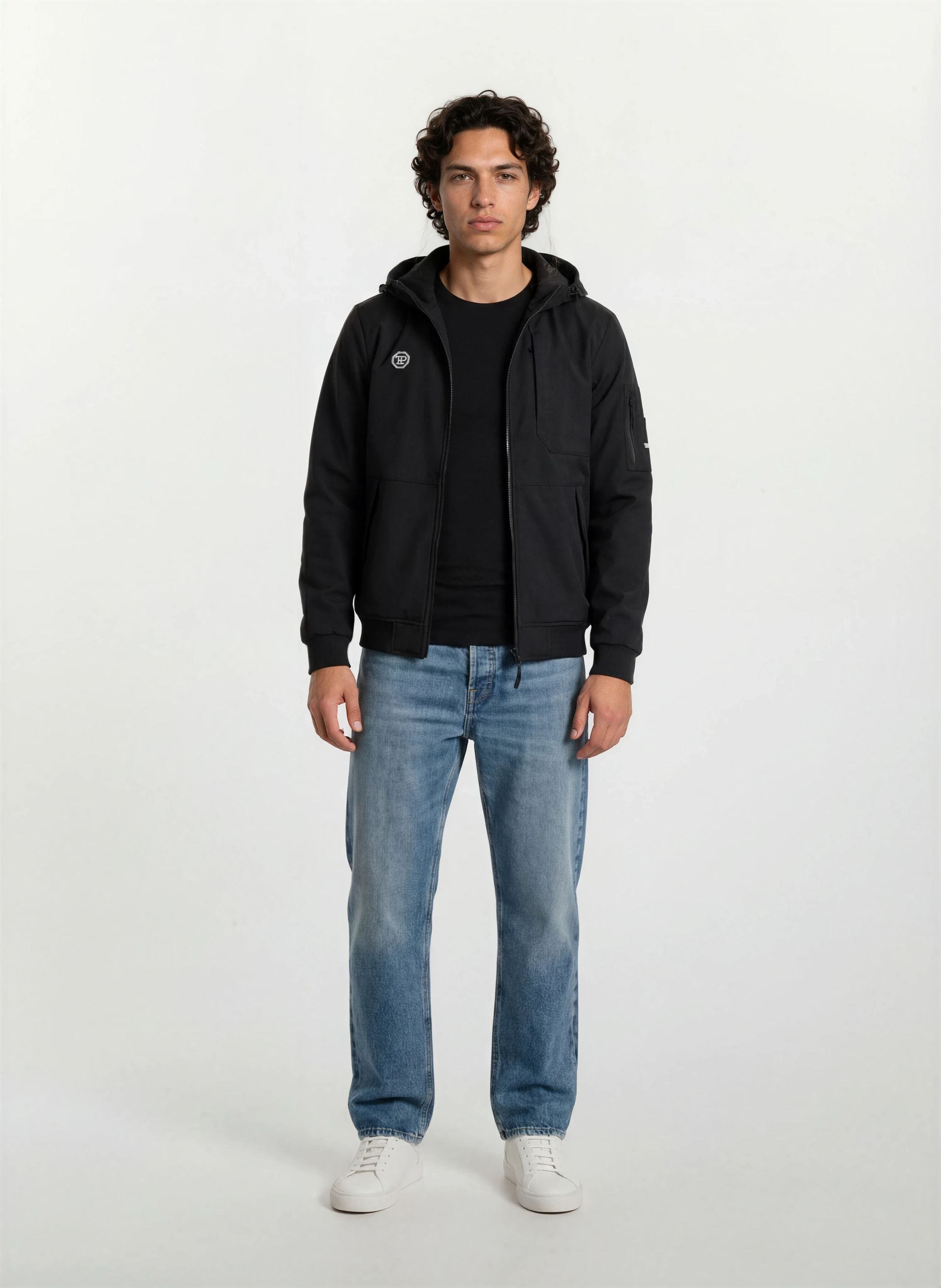 Ein Mann steht mit dem Gesicht nach vorne vor einem schlichten weißen Hintergrund. Er trägt eine Trueprodigy Leichte Herrenjacke mit Brust- und Ärmelbadge über einem schwarzen T-Shirt, blaue Jeans und weiße Turnschuhe.