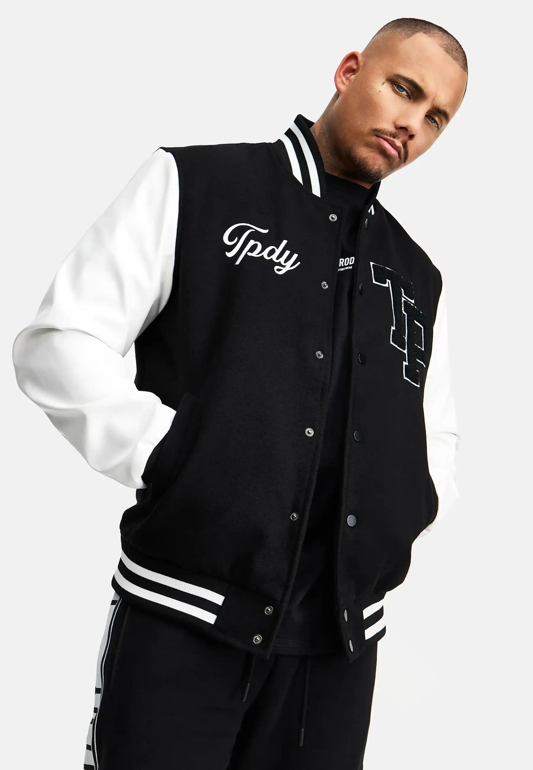 Ein Mann steht mit den Händen in den Taschen einer schwarz-weißen trueprodigy Chuck Retro Collage Jacke mit Logo, gestylt wie eine klassische College-Jacke mit gestickten Details. Er trägt eine schwarze Hose mit weißen Streifen und blickt in die Kamera.