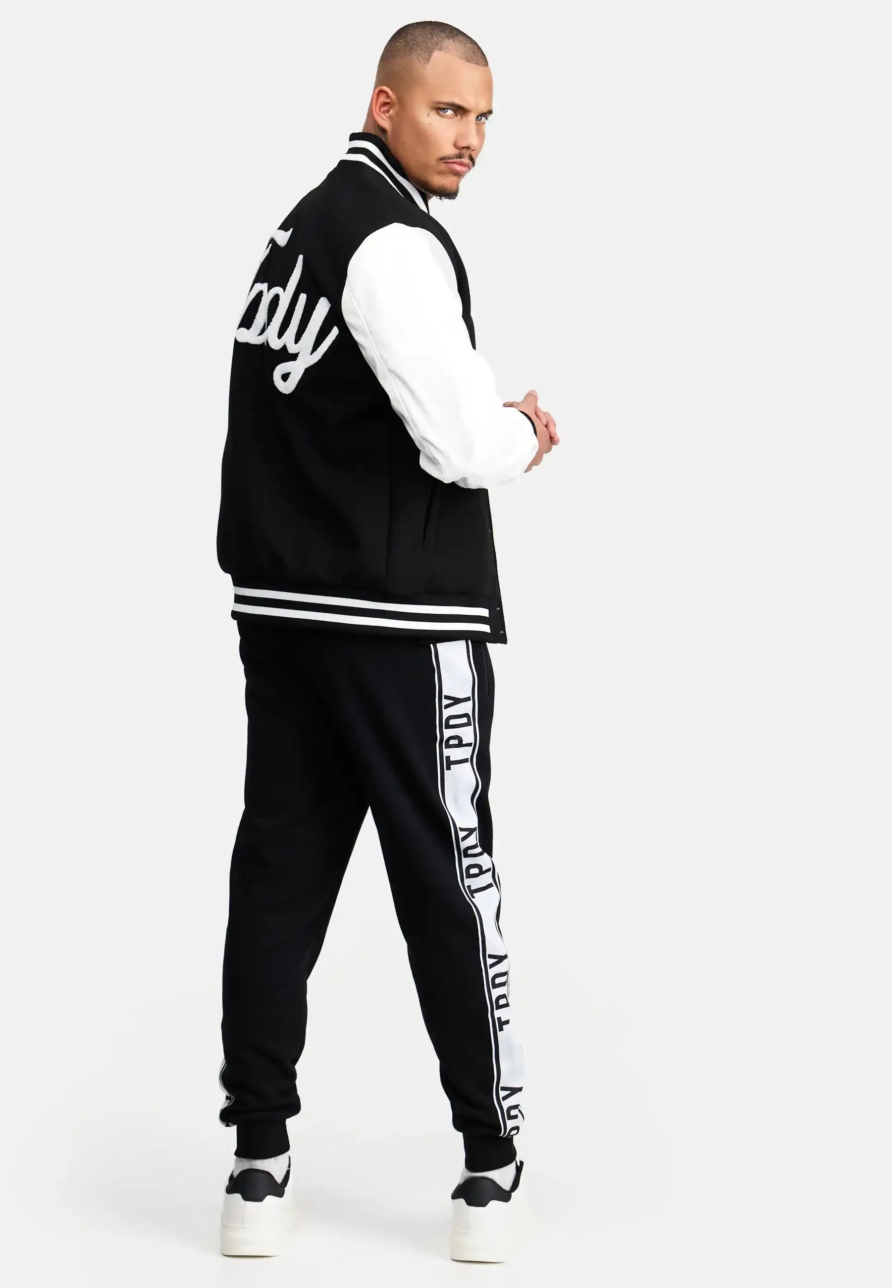 Ein Mann blickt nach hinten und wendet seinen Kopf der Kamera zu. Er trägt eine schwarz-weiße trueprodigy Chuck Retro Collage Jacke mit Logo, eine schwarze Trainingshose mit weißen Seitenstreifen und weiße Turnschuhe.