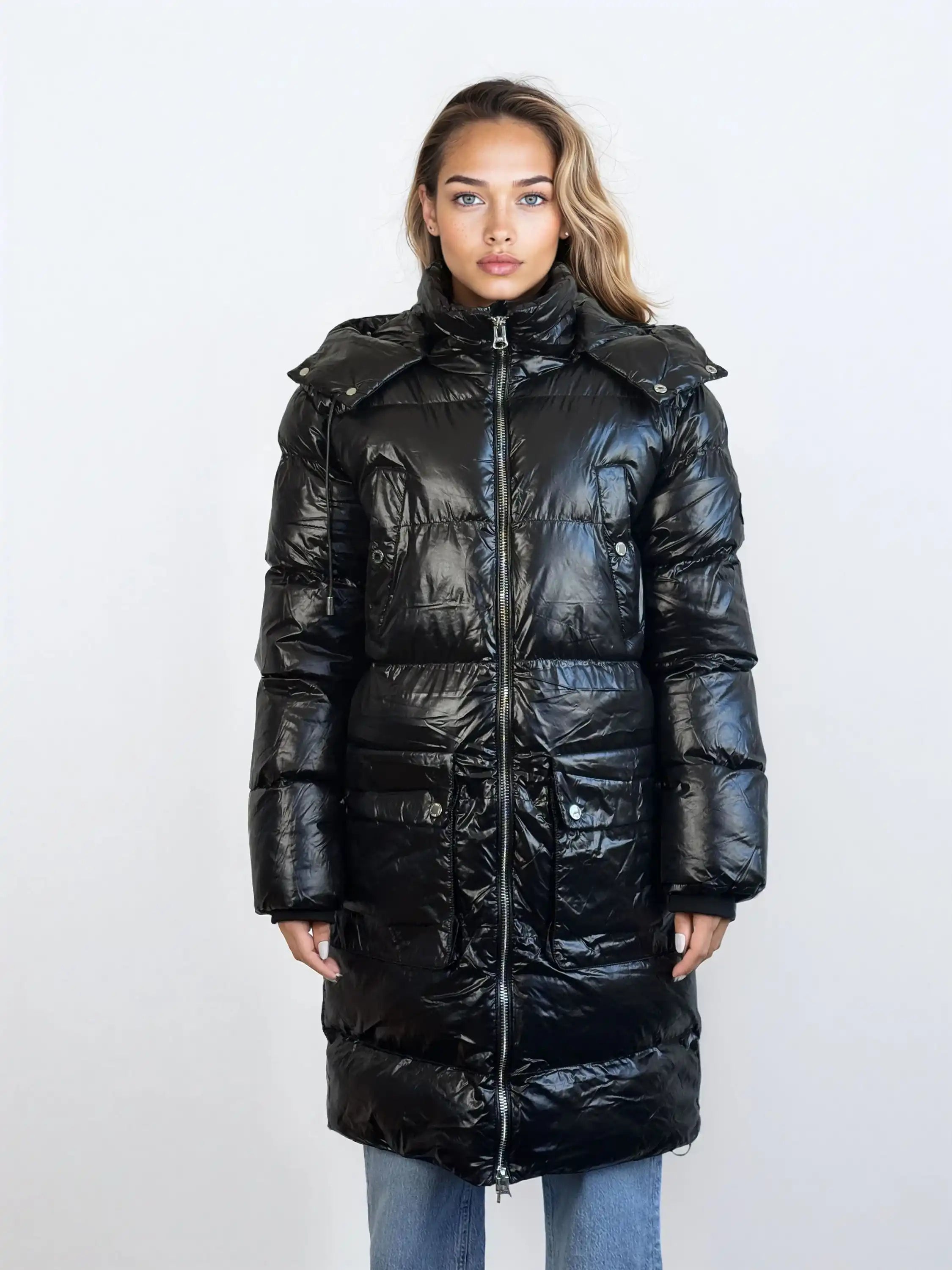 Eine Frau steht mit Blick nach vorne in einer glänzenden schwarzen trueprodigy Damen Winterjacke mit abnehmbarer Kapuze im Regular Fit mit Reißverschluss und hohem Kragen. Sie trägt eine blaue Jeans, die Hände an den Seiten, vor einem schlichten hellen Hintergrund.