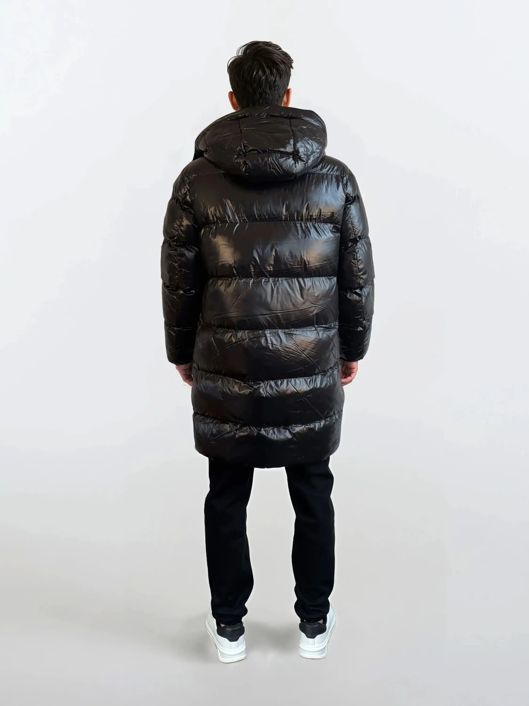 Eine Person mit kurzen dunklen Haaren schaut von der Kamera weg und trägt eine glänzende trueprodigy Herren Winterjacke mit abnehmbarer Kapuze im Regular Fit, eine schwarze Hose und weiße Turnschuhe vor einem schlichten hellen Hintergrund.
