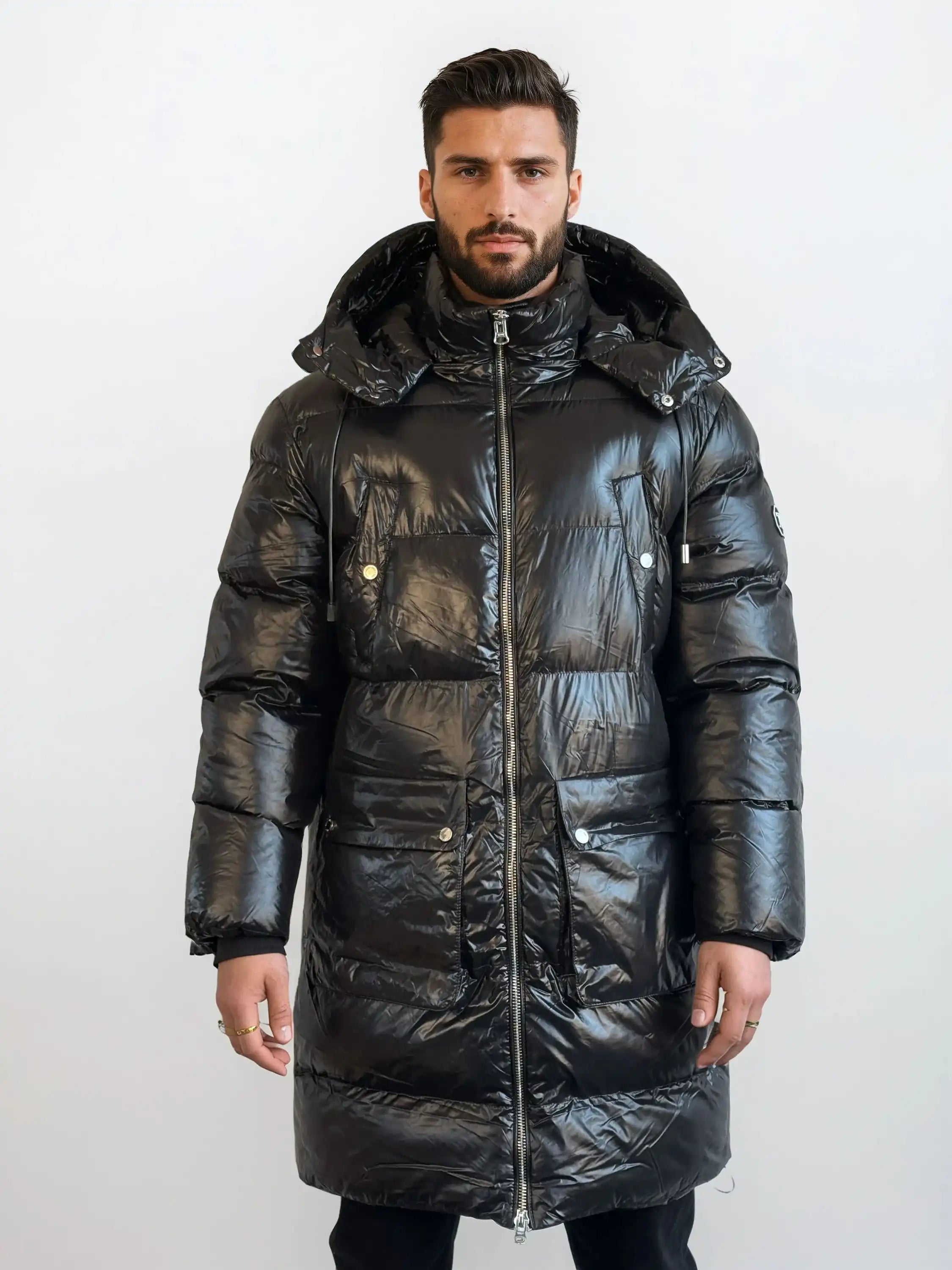 Ein Mann steht mit dem Gesicht nach vorne in einer glänzenden, langen schwarzen trueprodigy Herren Winterjacke mit abnehmbarer Kapuze im Regular Fit, die mit einem Frontreißverschluss und Druckknopftaschen ausgestattet ist, vor einem schlichten weißen Hintergrund.
