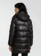 Eine Person mit dunklem, gewelltem Haar ist von hinten zu sehen, die eine glänzende trueprodigy Damen Winterjacke mit abnehmbarer Kapuze im Regular Fit trägt. Die glänzende Oberfläche der Jacke steht im Kontrast zu dem einfarbigen, hellen Hintergrund.